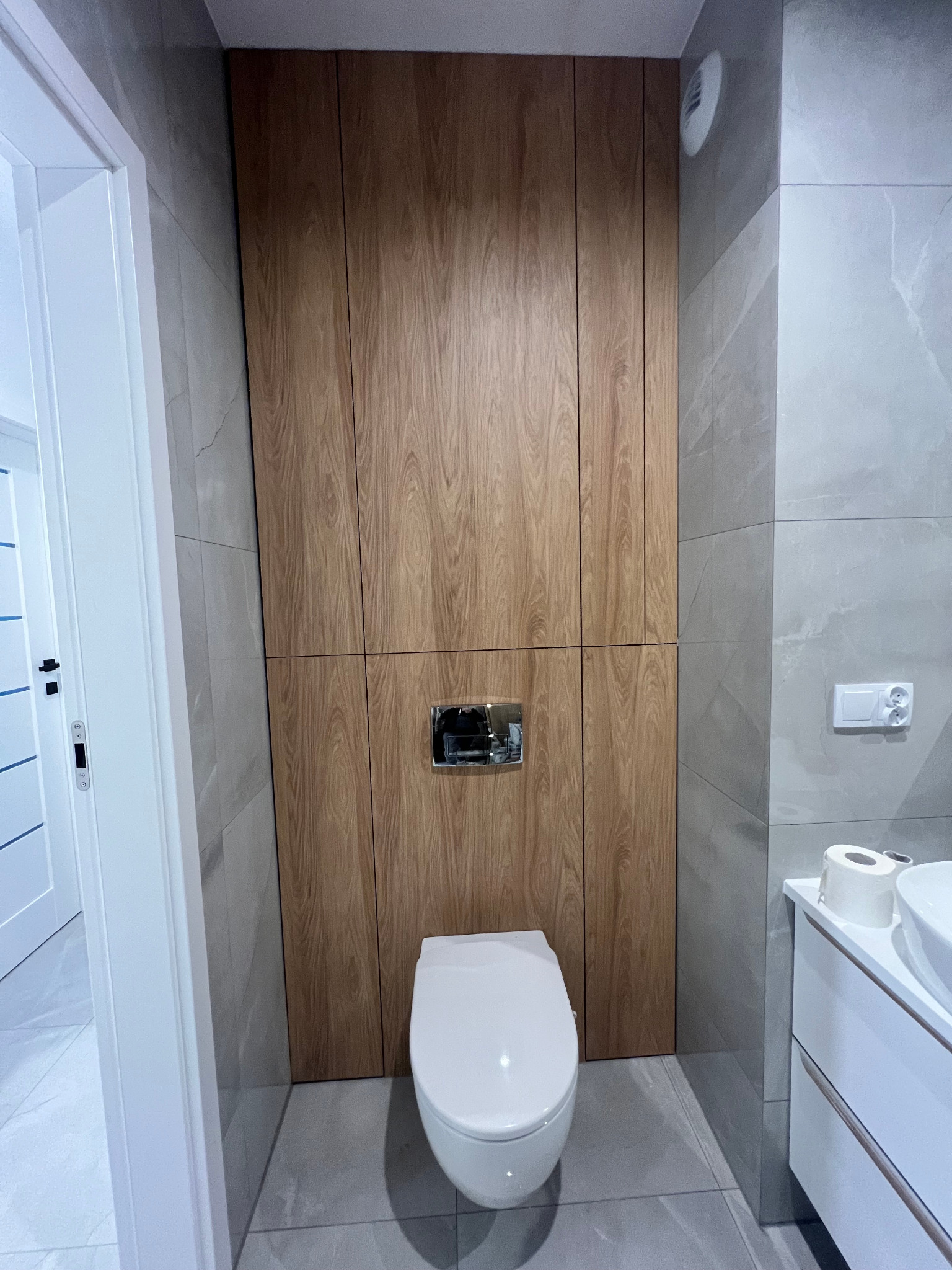 Nowoczesna łazienka z drewnianą zabudową nad toaletą, szarą glazurą i białą umywalką z szafką.