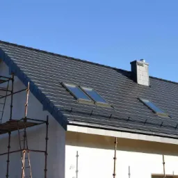 Nowy dach z ciemnej dachówki ceramicznej, z oknami połaciowymi i kominem z cegły, widoczny fragment elewacji i rusztowania.