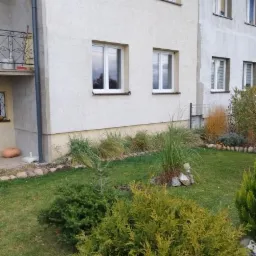 Dwupiętrowy budynek z białymi oknami i zadbanym ogrodem z krzewami iglastymi.