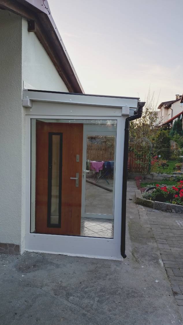 Biała rama okienna PCV z drewnianymi drzwiami wejściowymi, widok z zewnątrz, na podwórku suszarka z praniem, w tle ogród z czerwonymi kwiatami.