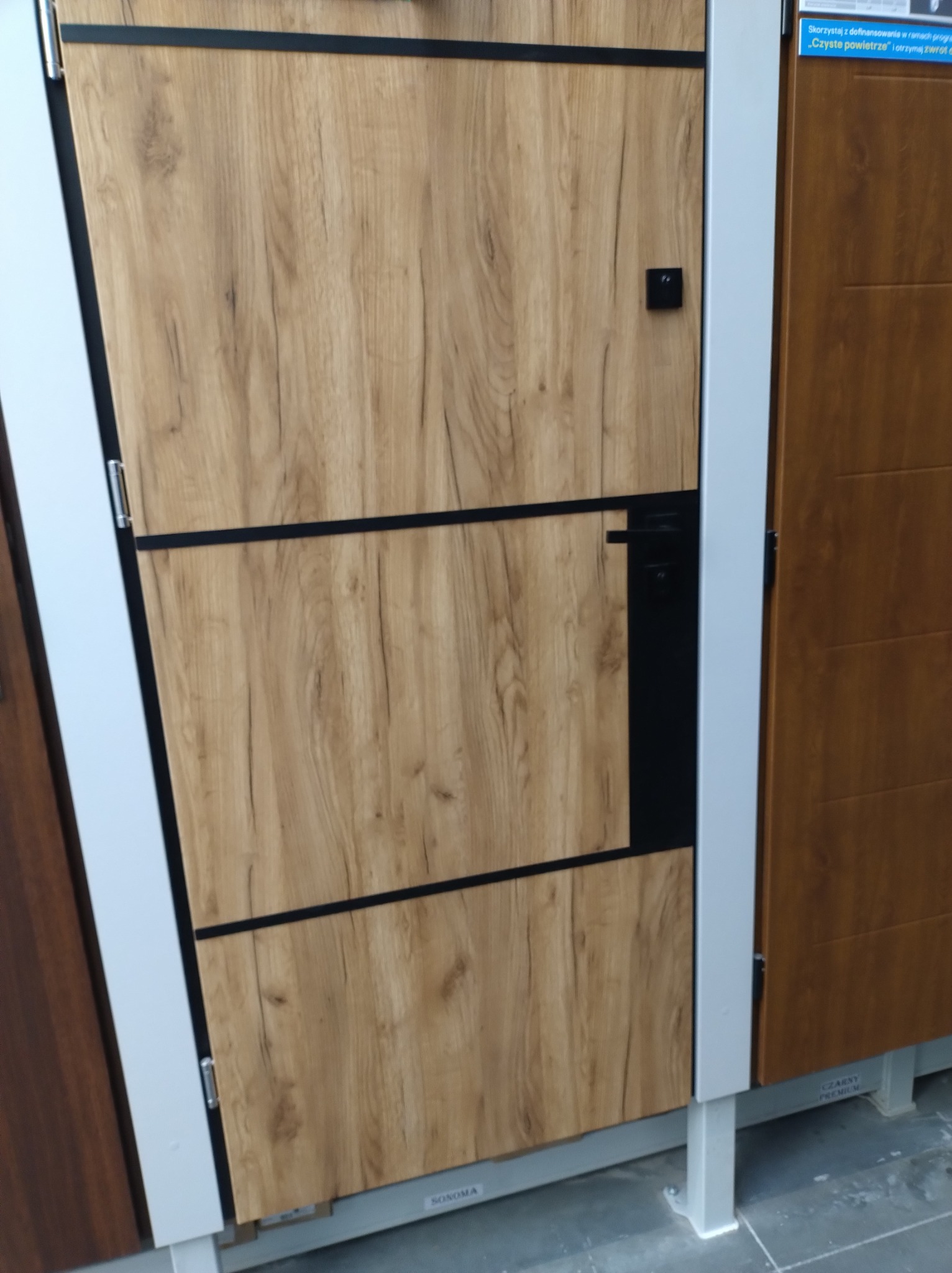 Prezentacja drzwi z okleiną drewnopodobną w odcieniach brązu, z czarnymi akcentami, widoczne okucie i fragment etykiety producenta.
