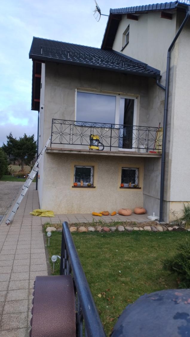 Dom z balkonem, na którym stoi żółty odkurzacz przemysłowy. Pod oknami leżą dynie, a obok ściany oparta jest drabina.