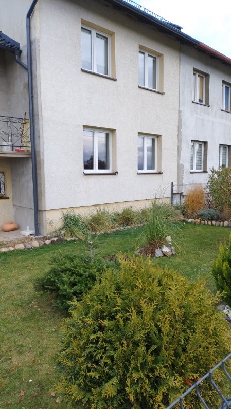 Dwupiętrowy budynek z białymi oknami i zadbanym ogrodem z krzewami iglastymi.