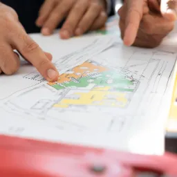 Dwie dłonie wskazują na kolorowy plan architektoniczny z naniesionymi wymiarami, obok leży poziomica