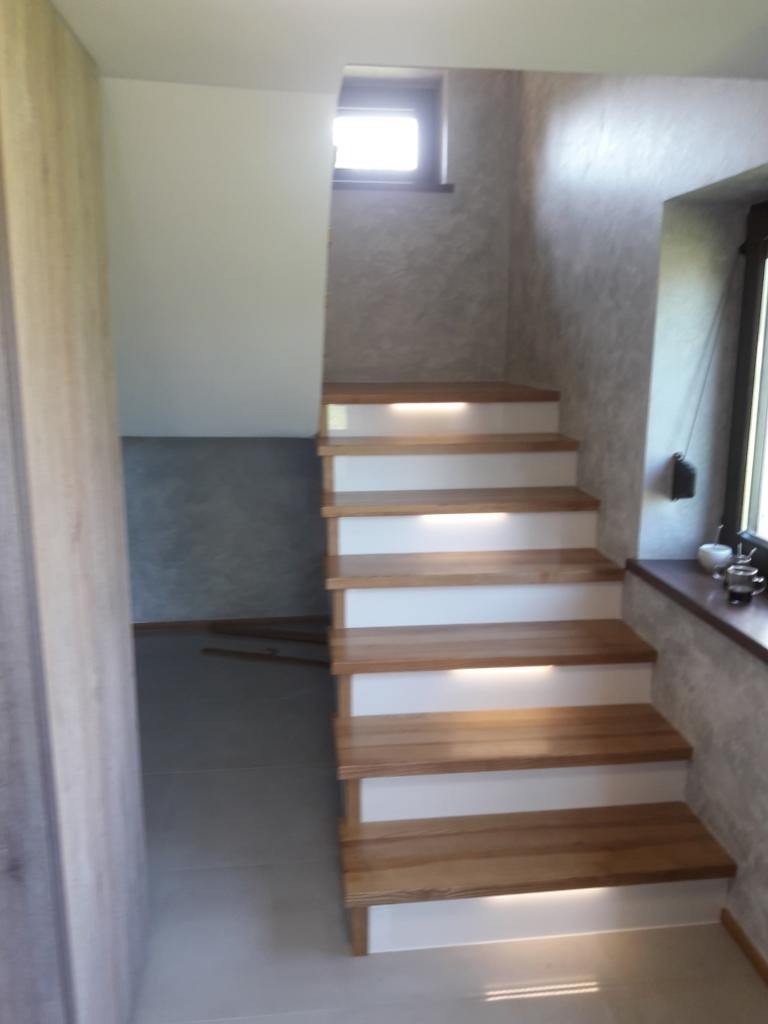 Nowoczesne schody drewniane z białymi podstopnicami i subtelnym oświetleniem LED, widok z dołu na tle ściany z dekoracyjną fakturą i oknem.