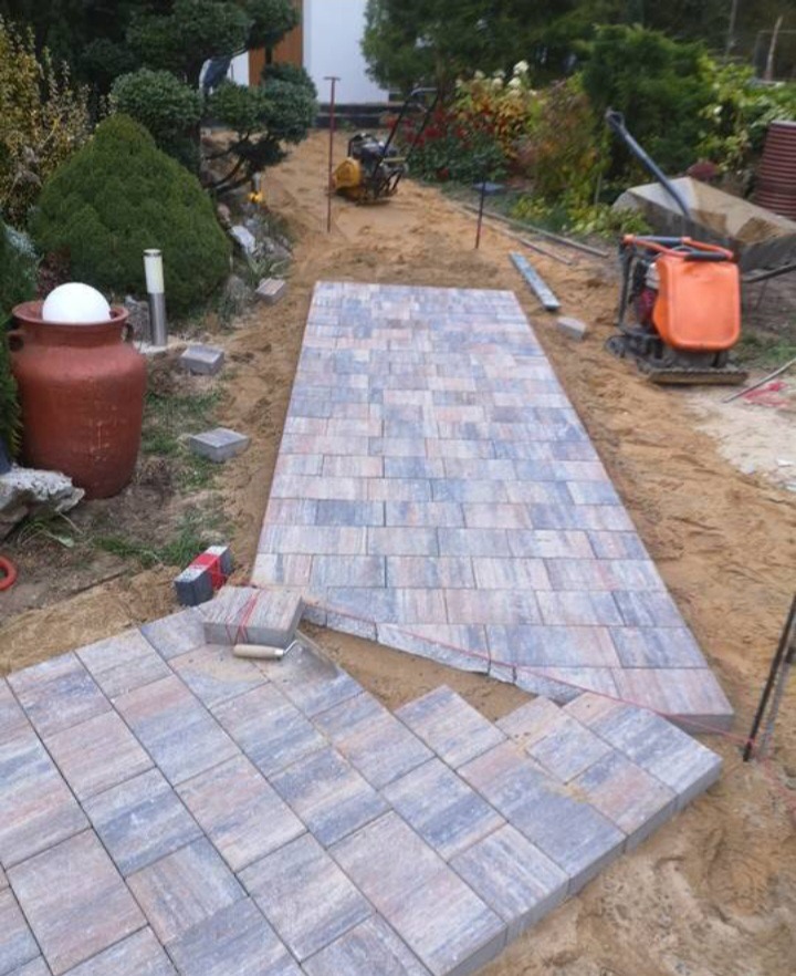 Ścieżka z kostki brukowej w trakcie układania, widoczna zagęszczarka i narzędzia brukarskie, elementy ogrodu w tle.