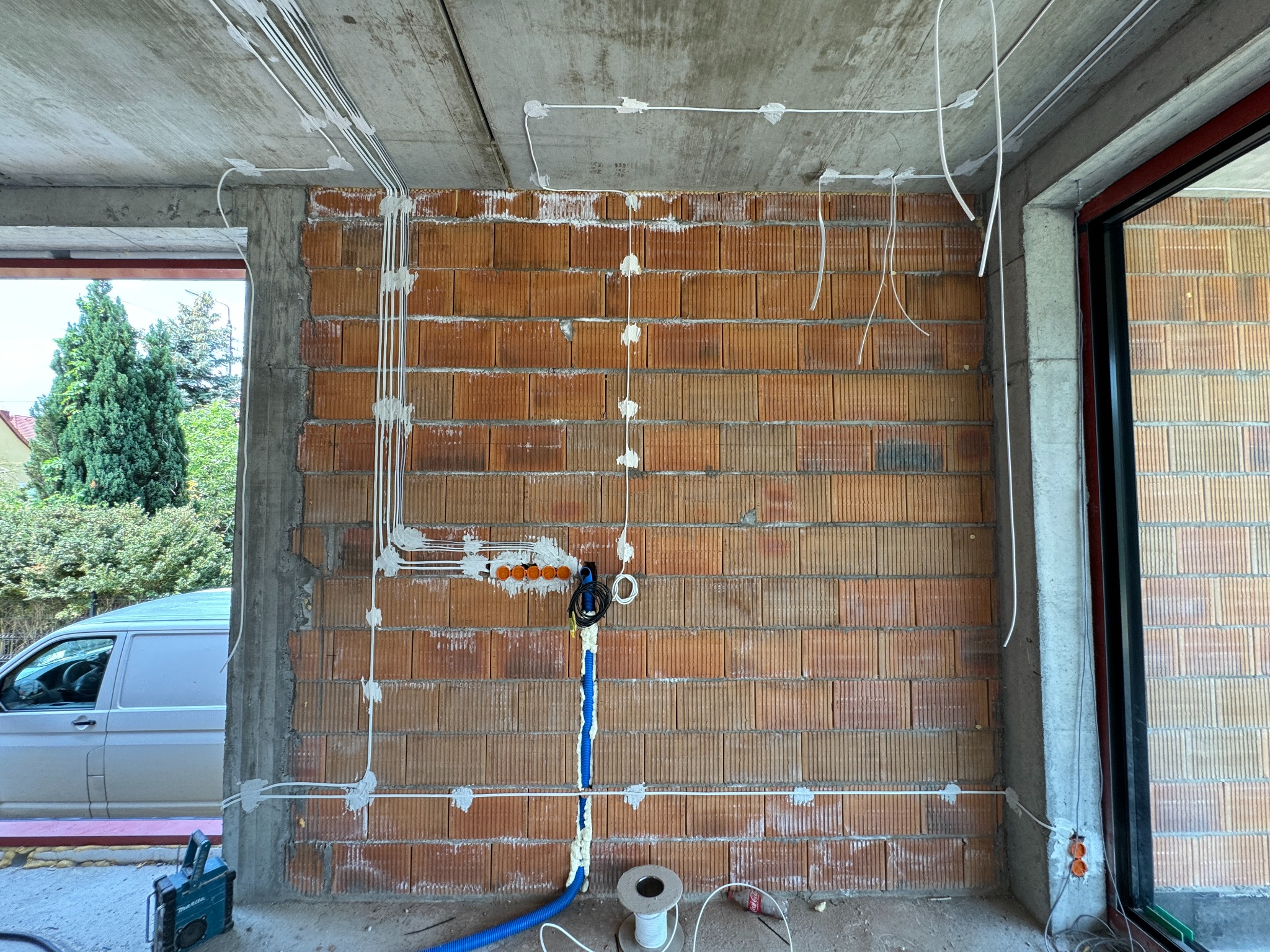 Surowa ściana z cegły dziurawki w trakcie montażu instalacji elektrycznej: białe kable przymocowane do ściany za pomocą szarego gipsu, niebieski peszel z kablami w pianie montażowej, puszki...