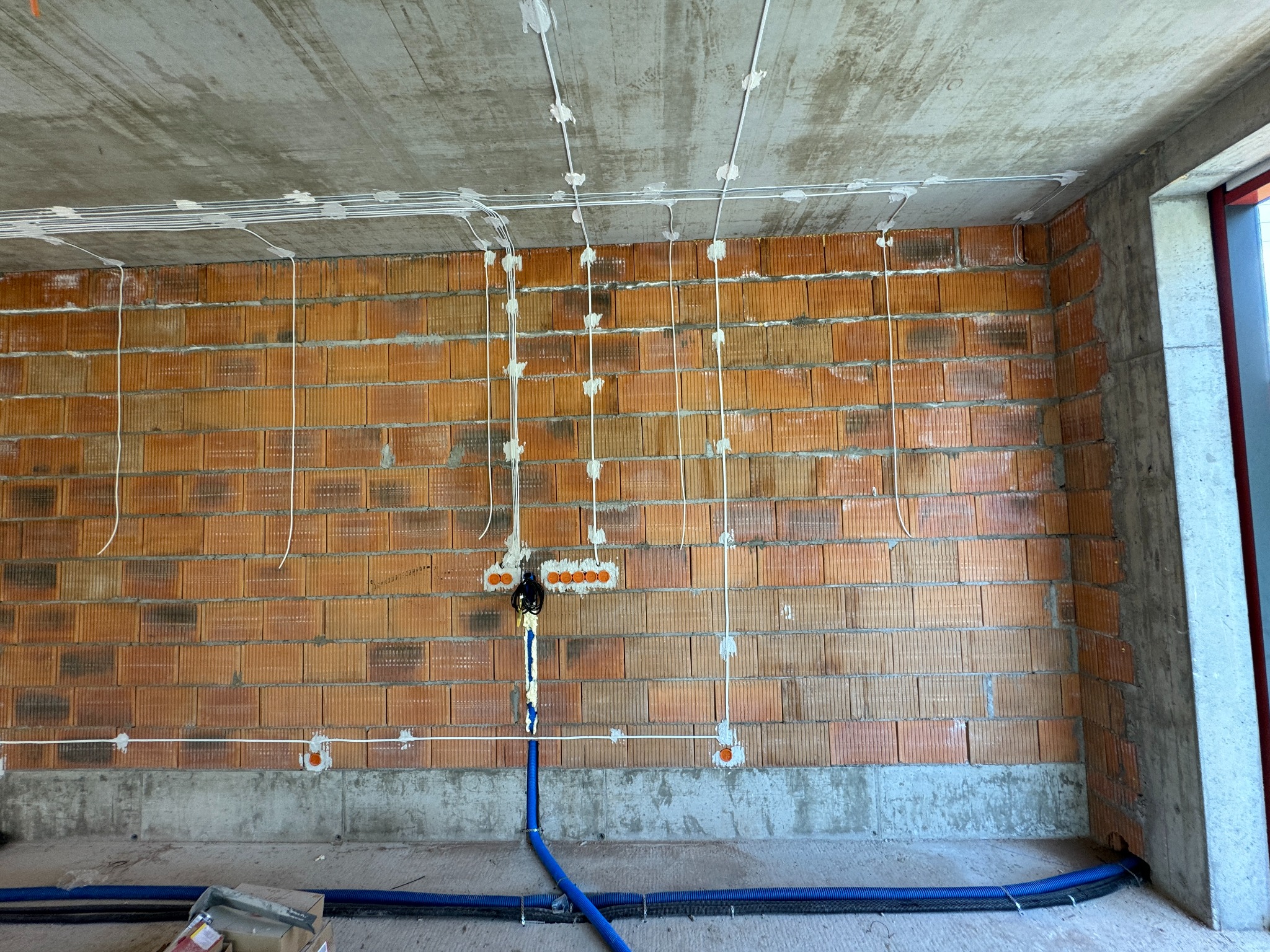 Wewnętrzna instalacja elektryczna w trakcie montażu: przewody prowadzone po ceglanej ścianie i betonowym suficie, zabezpieczone uchwytami, widoczne puszki instalacyjne i rury karbowane w kolorze...