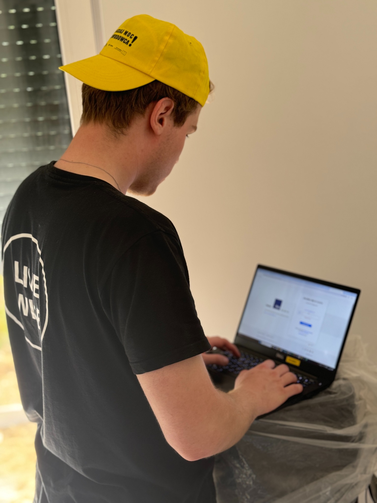 Mężczyzna w czarnej koszulce i żółtej czapce z daszkiem, tyłem do kamery, pracuje na czarnym laptopie postawionym na czymś owiniętym folią, prawdopodobnie podczas konfiguracji systemu.