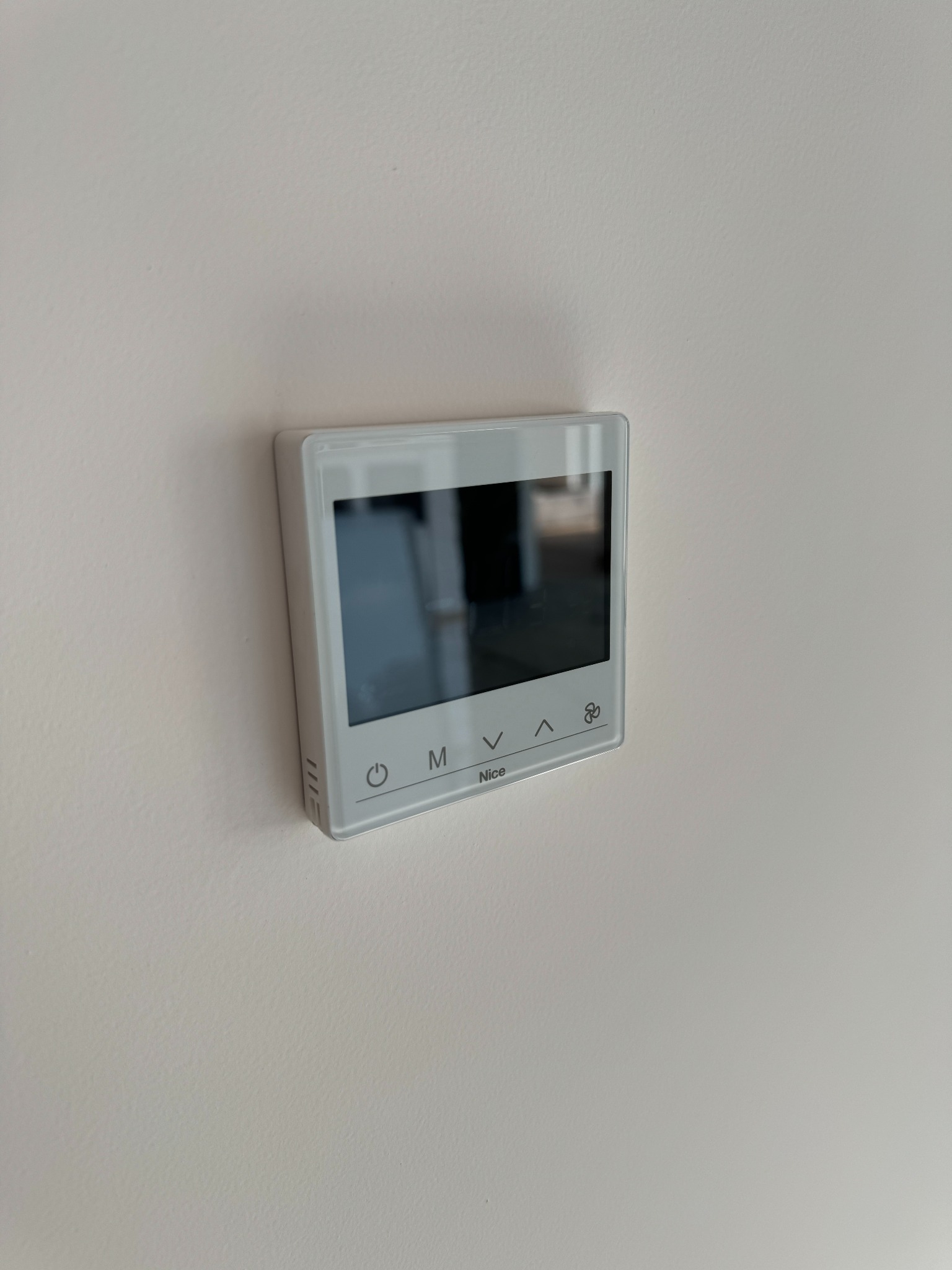 Biały panel sterowania z dotykowym ekranem, zamontowany na jasnej ścianie, widoczny z boku, z logo 'Nice' w dolnej części.