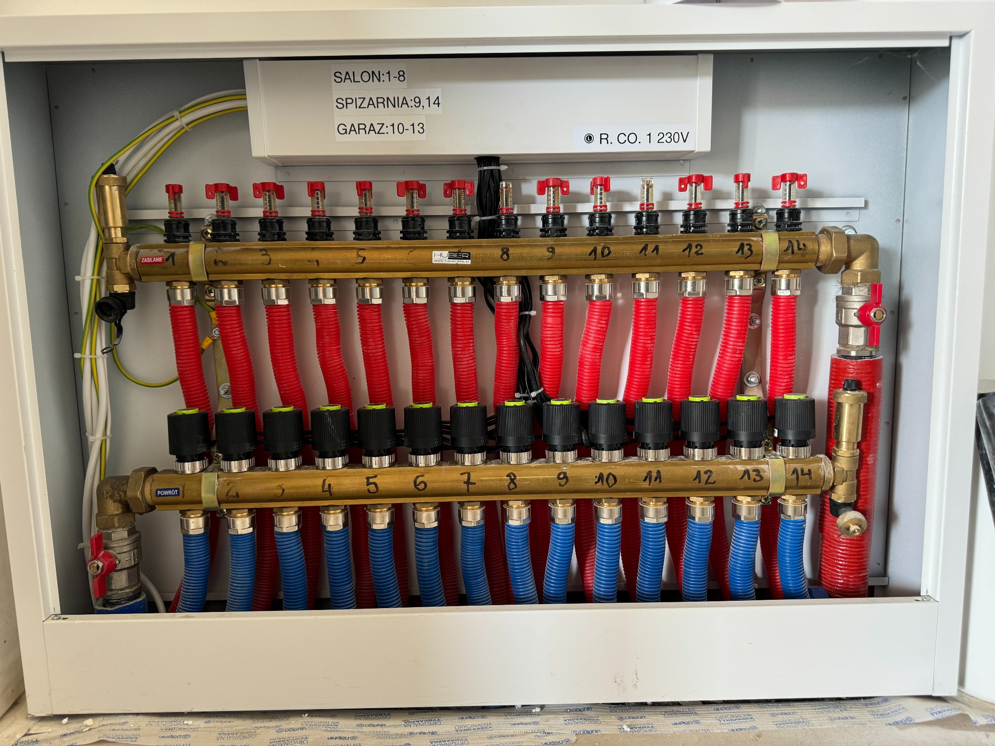 Rozdzielacz ogrzewania podłogowego w szafce, z numerowanymi zaworami i rurami w kolorach czerwonym i niebieskim, oznaczony opisami pomieszczeń: salon, spiżarnia, garaż.