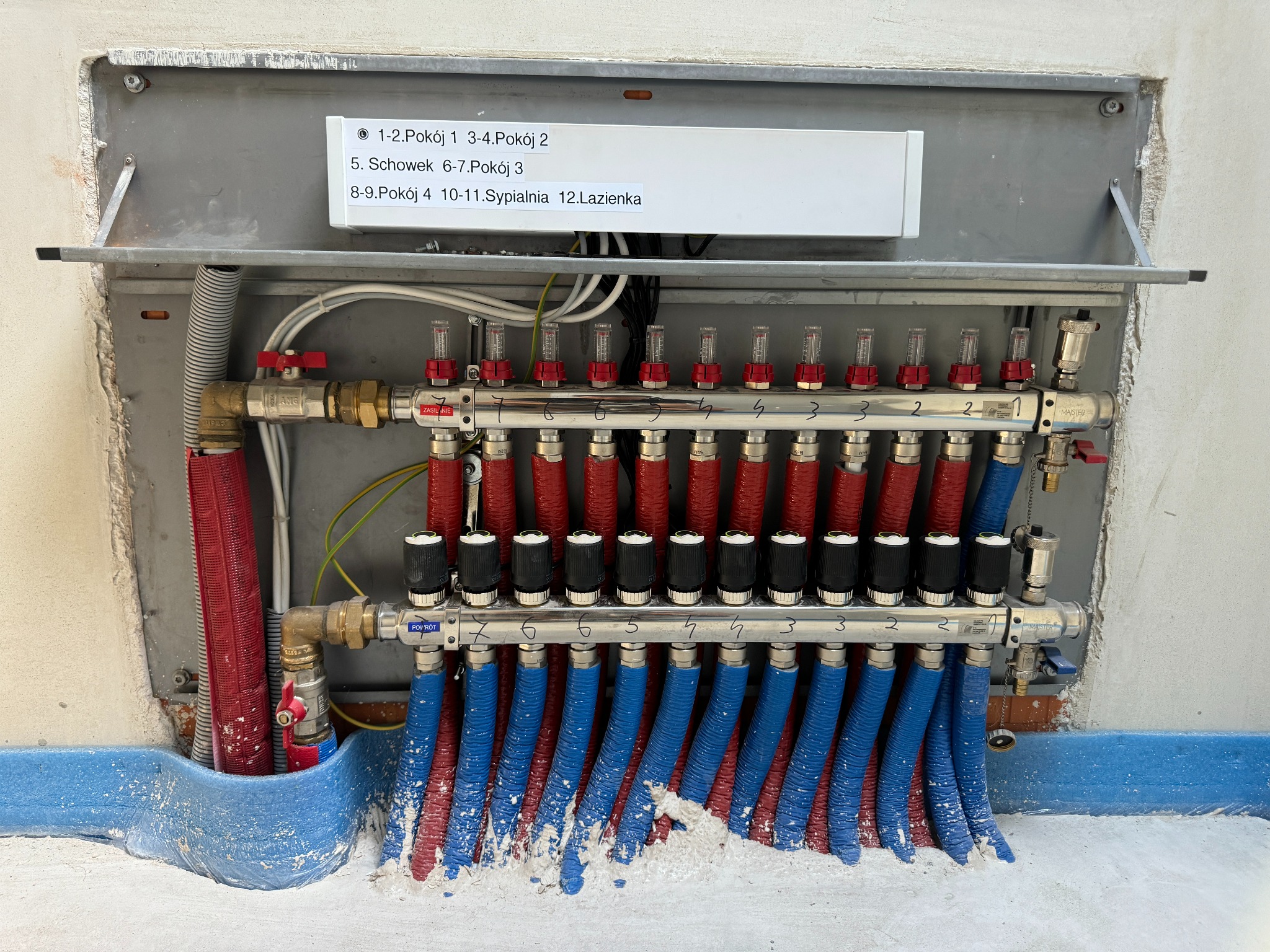 Rozdzielacz ogrzewania podłogowego z rurami w kolorze czerwonym i niebieskim, zamontowany na szarej płycie z etykietą opisującą obwody grzewcze dla różnych pomieszczeń, widoczne niedociągnięcia...