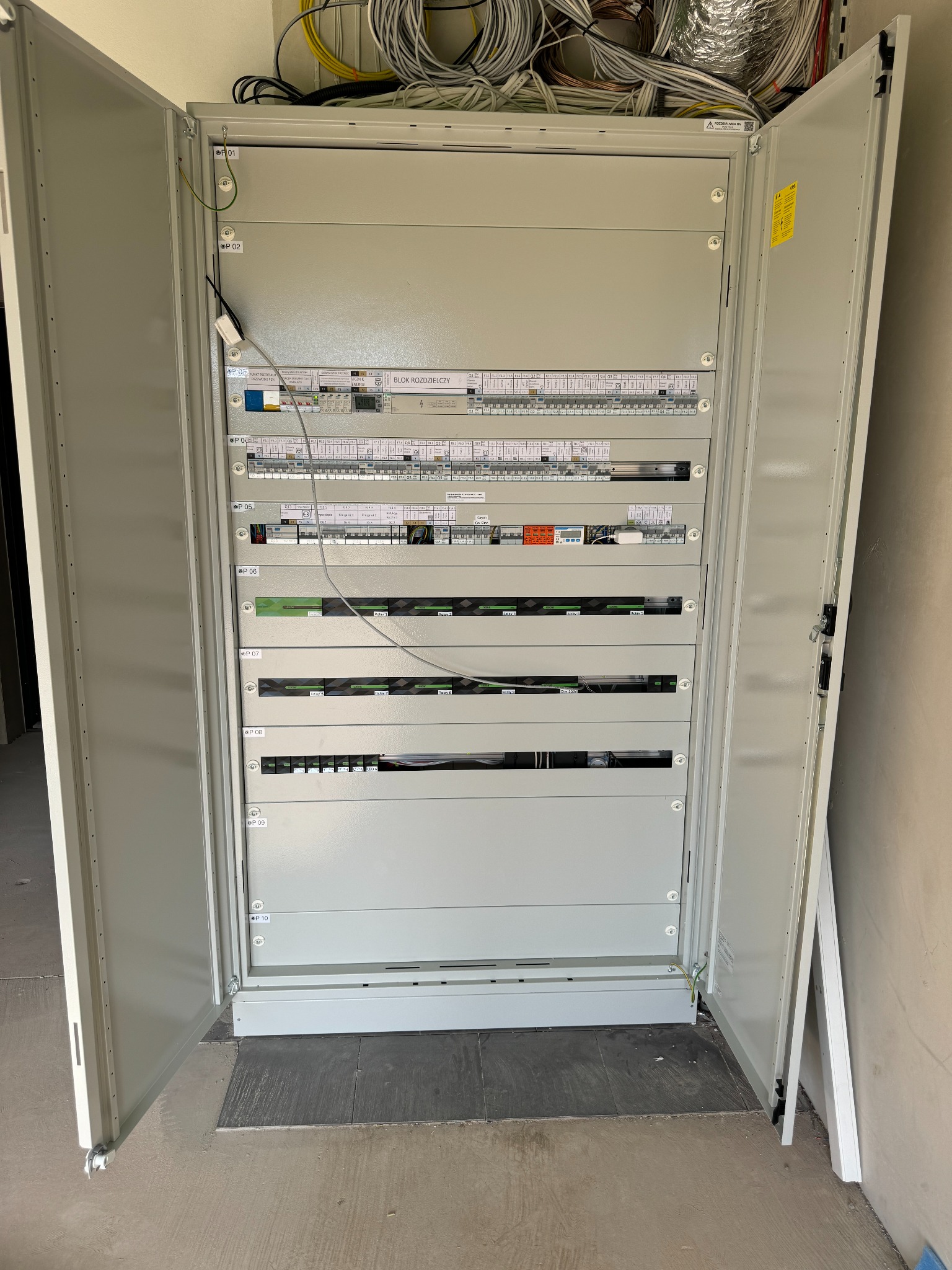 Otwarta szafa rozdzielcza z widocznymi zabezpieczeniami, bezpiecznikami i okablowaniem, zamontowana w nowym budynku z surowym wykończeniem ścian i podłogi.