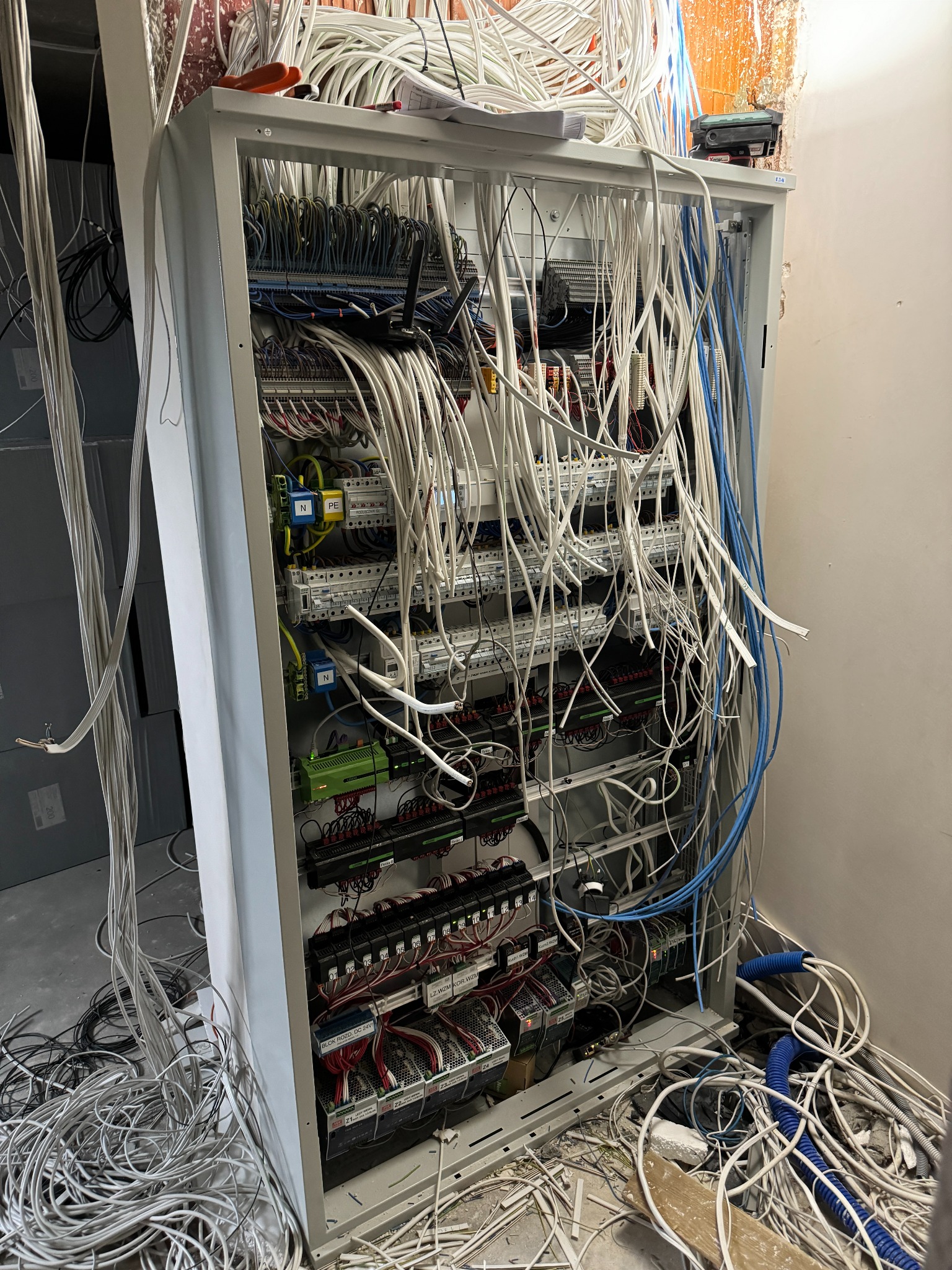 Otwarta szafa sterownicza w trakcie montażu, widoczne liczne kable białe i niebieskie, część z nich zwisa z sufitu i leży na podłodze, kable podłączone do modułów sterujących i zasilaczy wewnątrz...