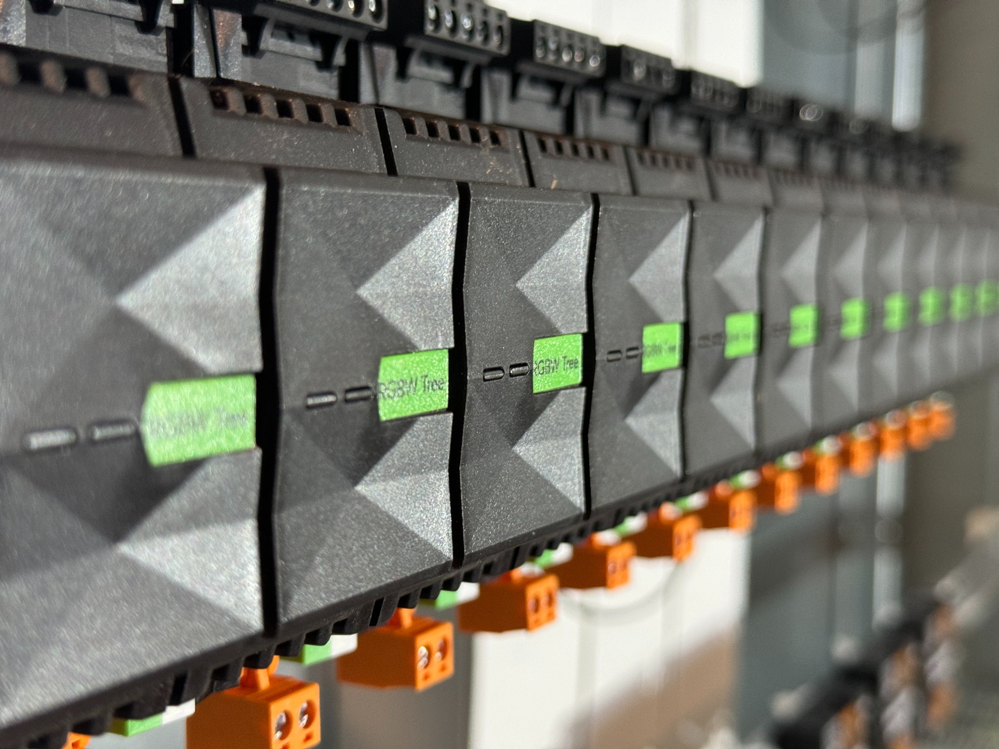Szereg modułów sterujących z zielonymi etykietami RGBW Tree i pomarańczowymi złączkami na szynie montażowej w rozdzielnicy elektrycznej.