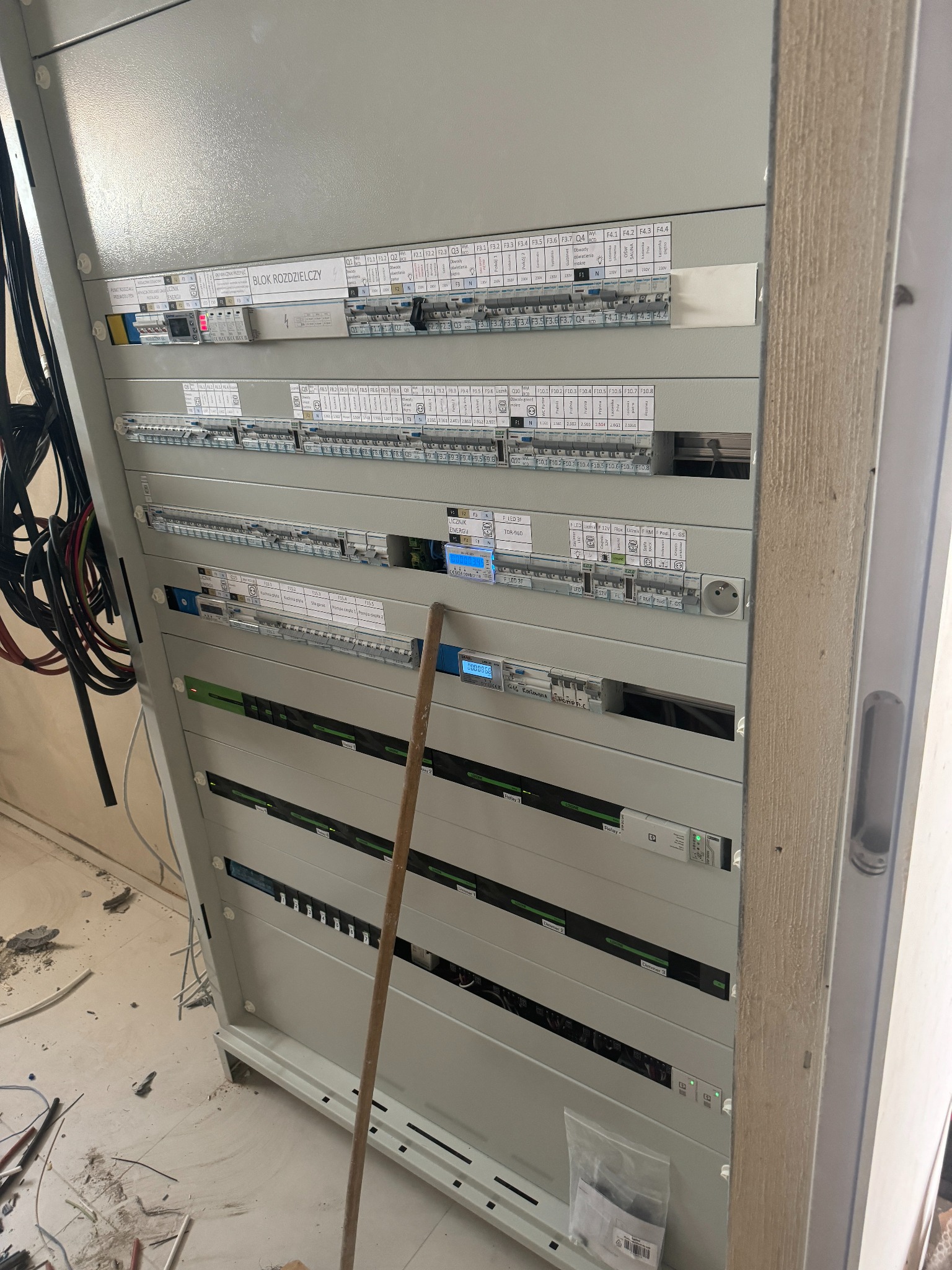 Szara szafa rozdzielcza z licznymi bezpiecznikami, etykietami i wyświetlaczami, widoczne kable i akcesoria na podłodze, co sugeruje trwające prace.