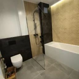 Nowoczesna łazienka po remoncie z kabiną prysznicową typu walk-in, podwieszaną toaletą i drewnopodobnymi płytkami ułożonymi w jodełkę na ścianie, widoczny otwarty schowek pod wanną.