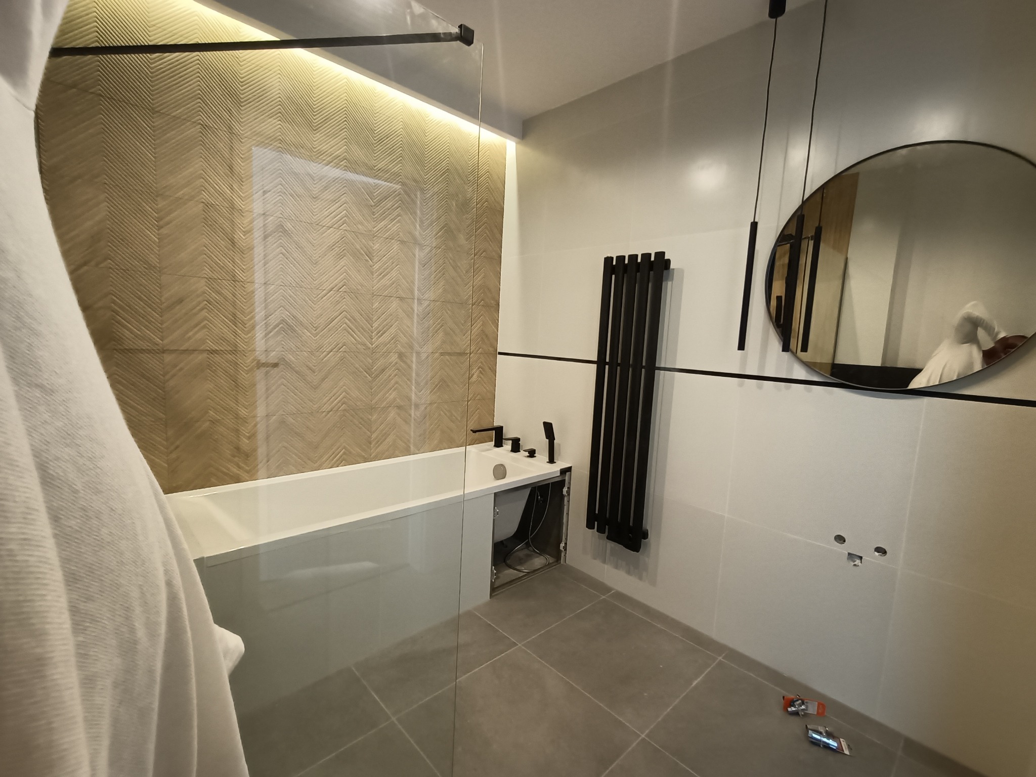 Nowoczesna łazienka w trakcie wykańczania: wanna z szybą, grzejnik drabinkowy, okrągłe lustro i płytki z wzorem jodełki, widoczne narzędzia na podłodze.