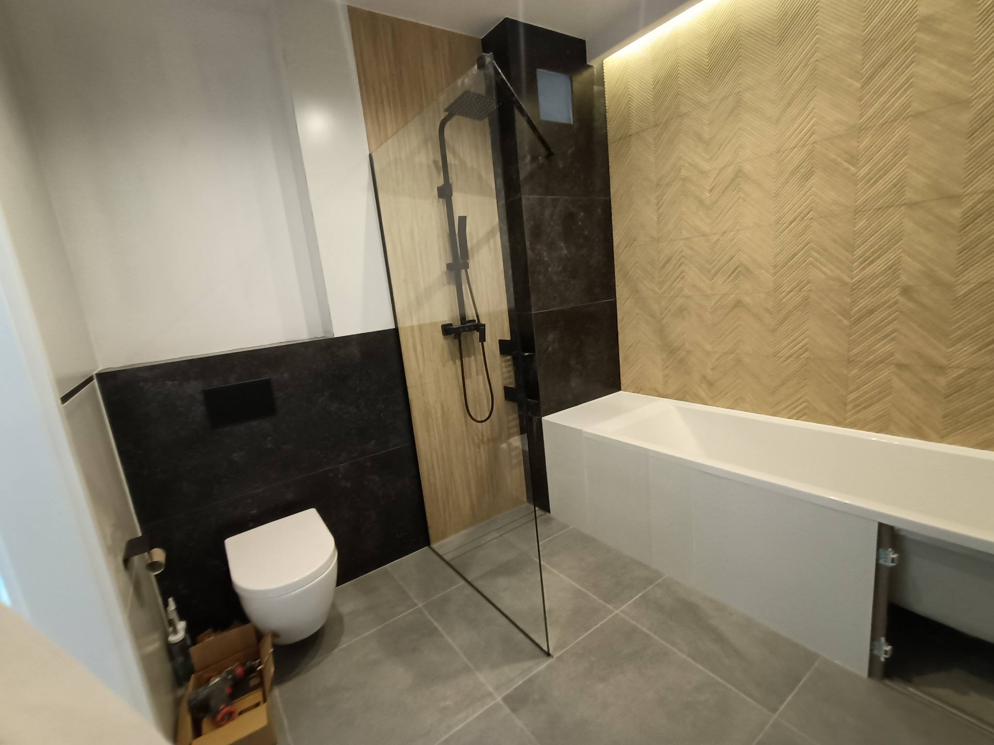 Nowoczesna łazienka po remoncie z kabiną prysznicową typu walk-in, podwieszaną toaletą i drewnopodobnymi płytkami ułożonymi w jodełkę na ścianie, widoczny otwarty schowek pod wanną.