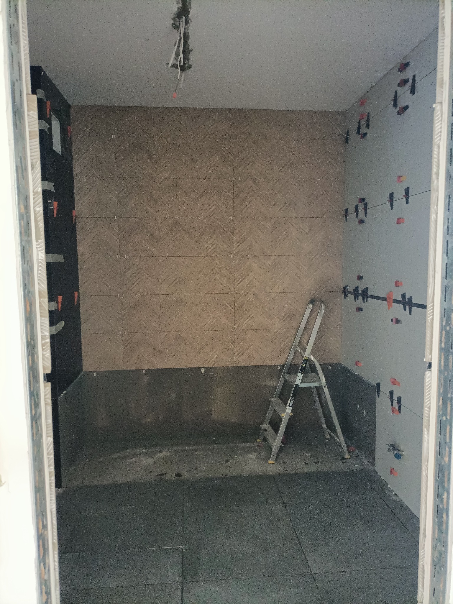 Łazienka w trakcie wykańczania: ułożone płytki w jodełkę na ścianie, czarne płytki częściowo ułożone na ścianie bocznej, drabina aluminiowa wewnątrz, system poziomowania płytek na ścianie bocznej.