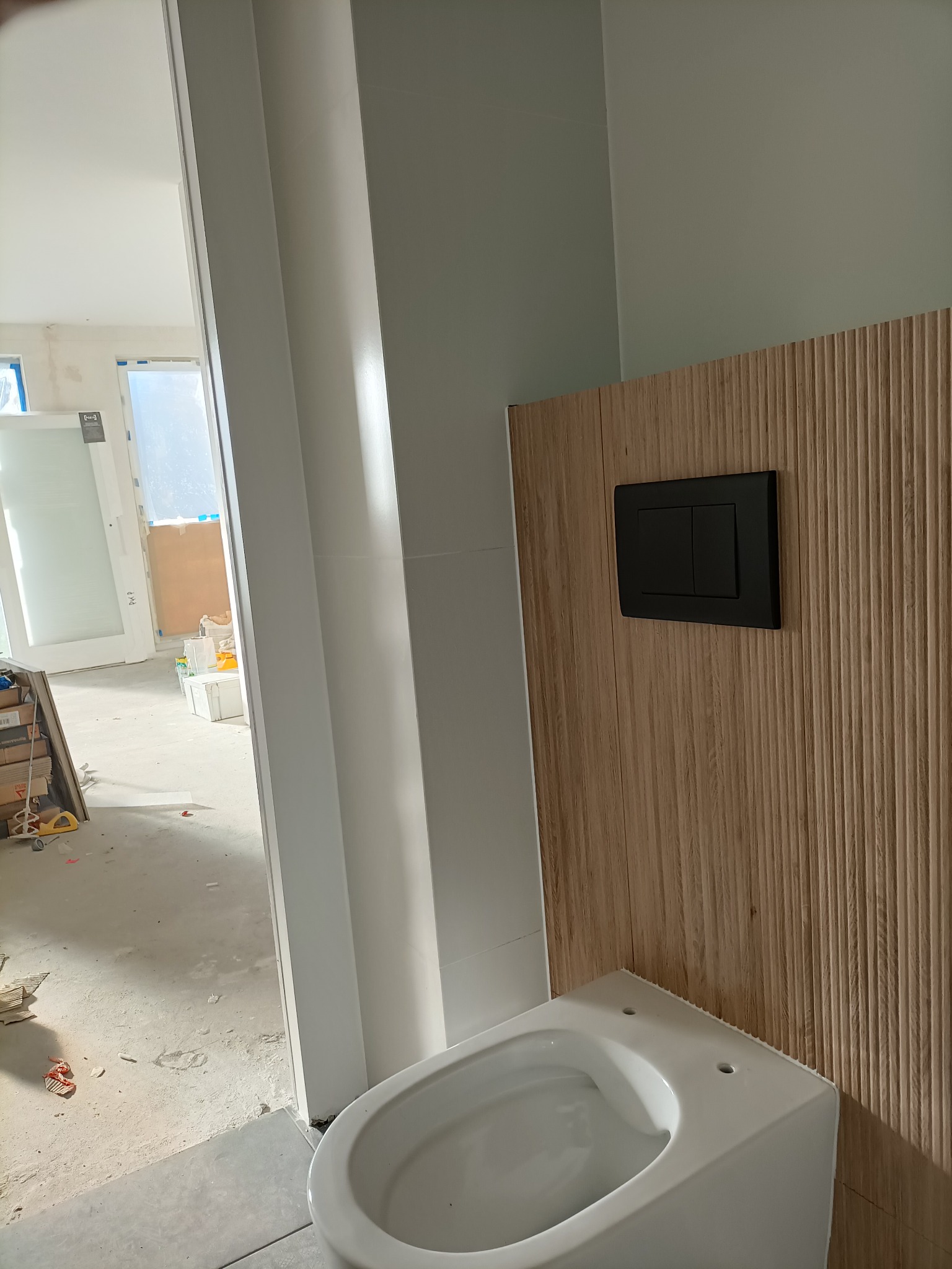 Montaż podtynkowego stelaża WC z drewnopodobną okładziną i czarnym przyciskiem spłukującym, widoczne przygotowania do wykończenia łazienki w surowym stanie.