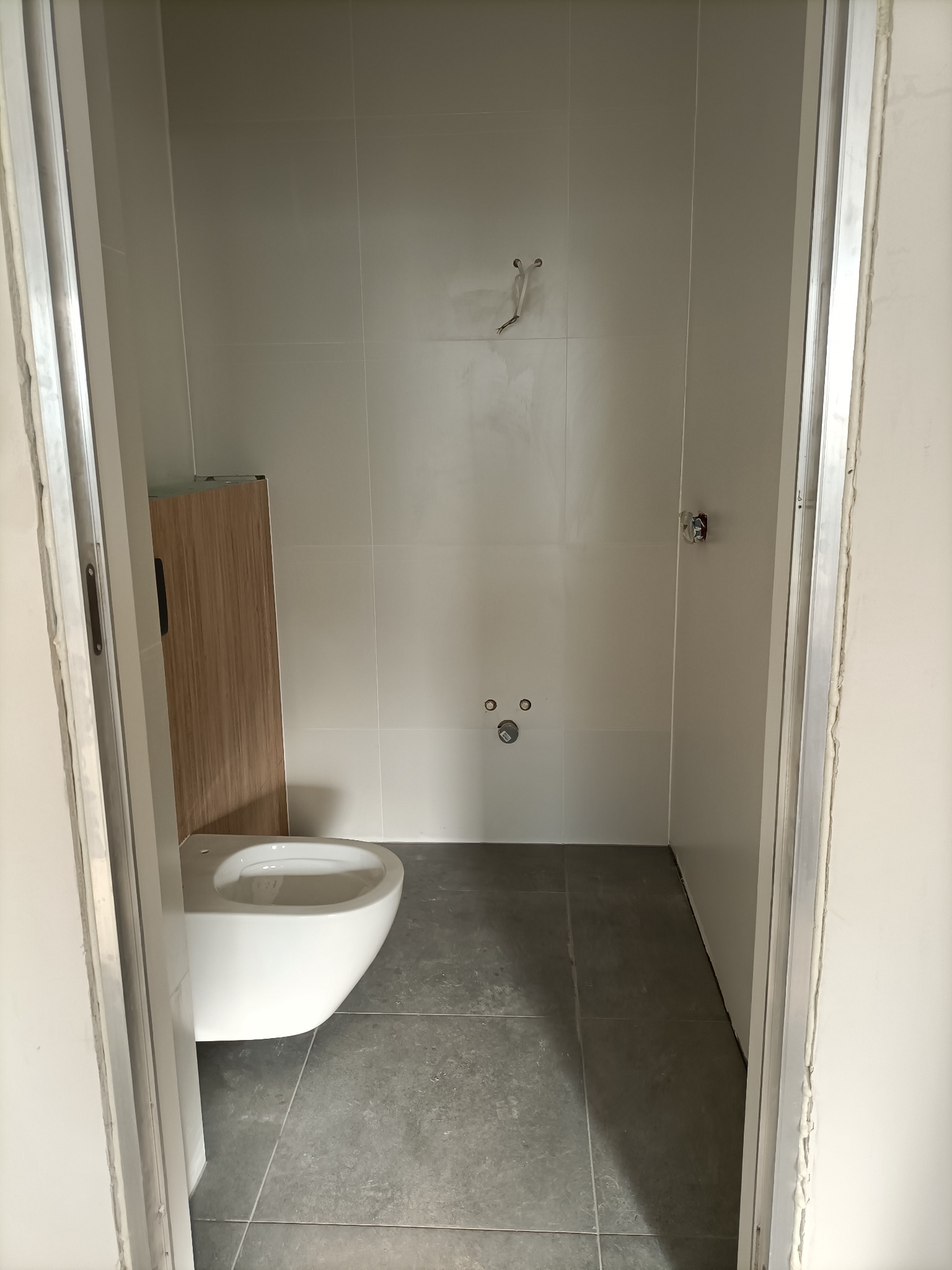 Wnętrze łazienki w trakcie wykańczania, widoczna podwieszana toaleta z zabudową, szare płytki podłogowe, białe płytki ścienne, wystające kable i niezamaskowane rury.