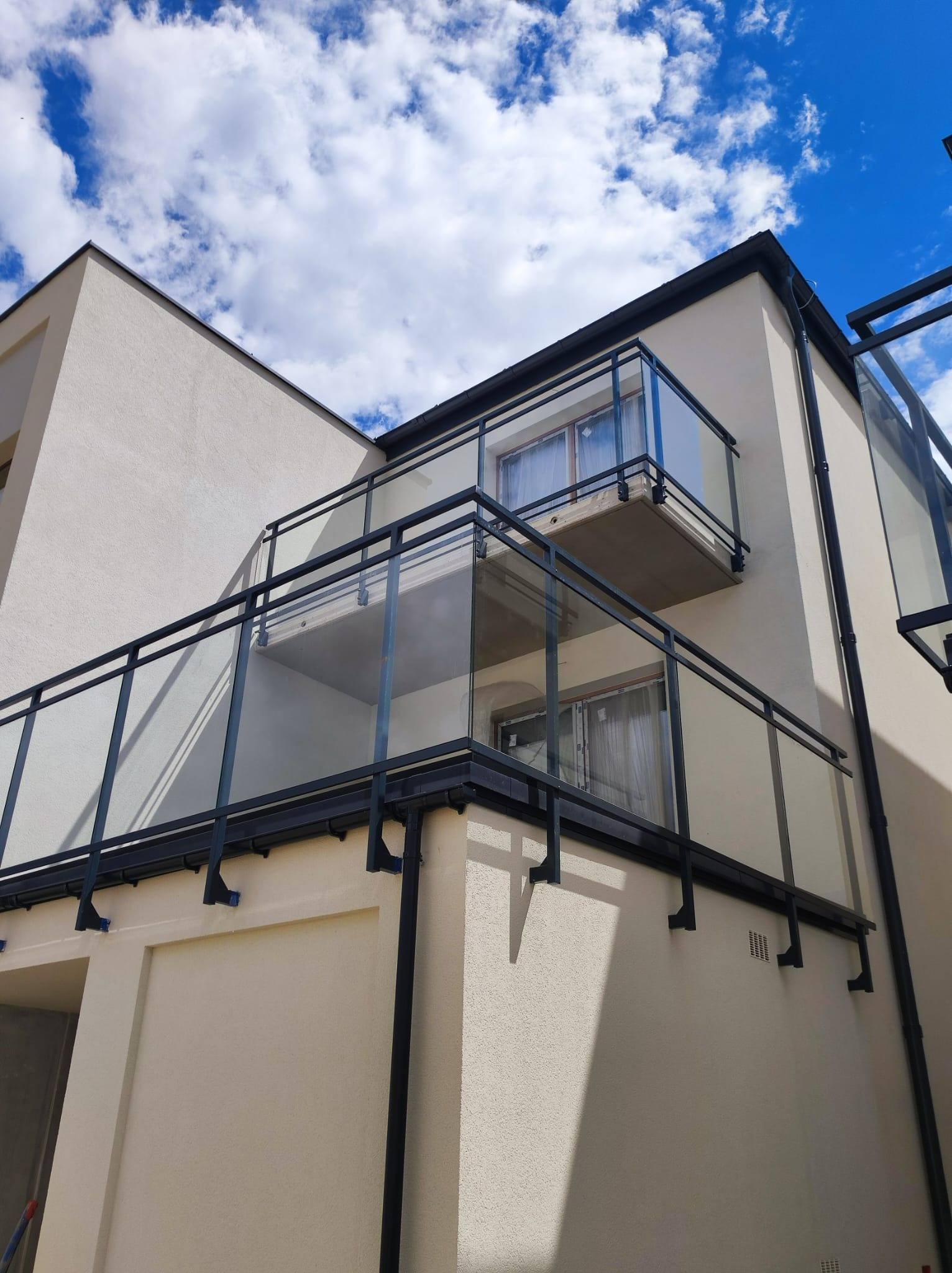 Nowoczesny budynek z balkonami zabezpieczonymi czarnymi ramami i szklanymi panelami, widok z dołu na tle błękitnego nieba z chmurami.