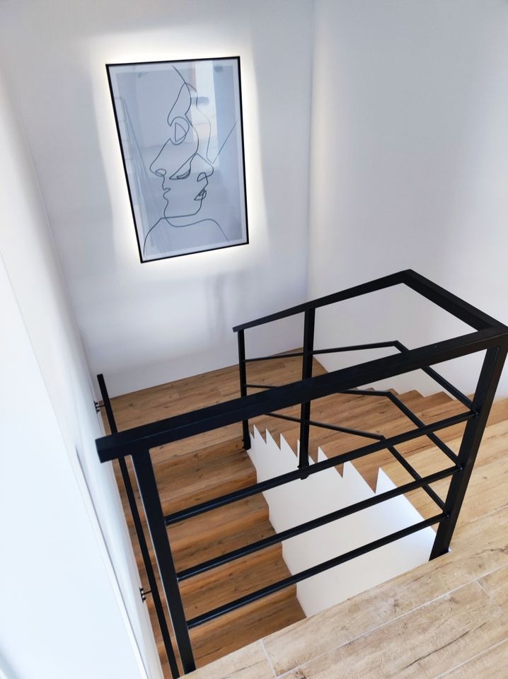 Nowoczesne schody z drewnianymi stopniami i czarną metalową balustradą, oświetlenie LED na ścianie z grafiką.