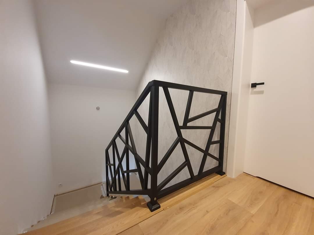 Czarna balustrada schodowa o geometrycznym wzorze, zamontowana w jasnym wnętrzu z drewnianą podłogą i białymi ścianami.