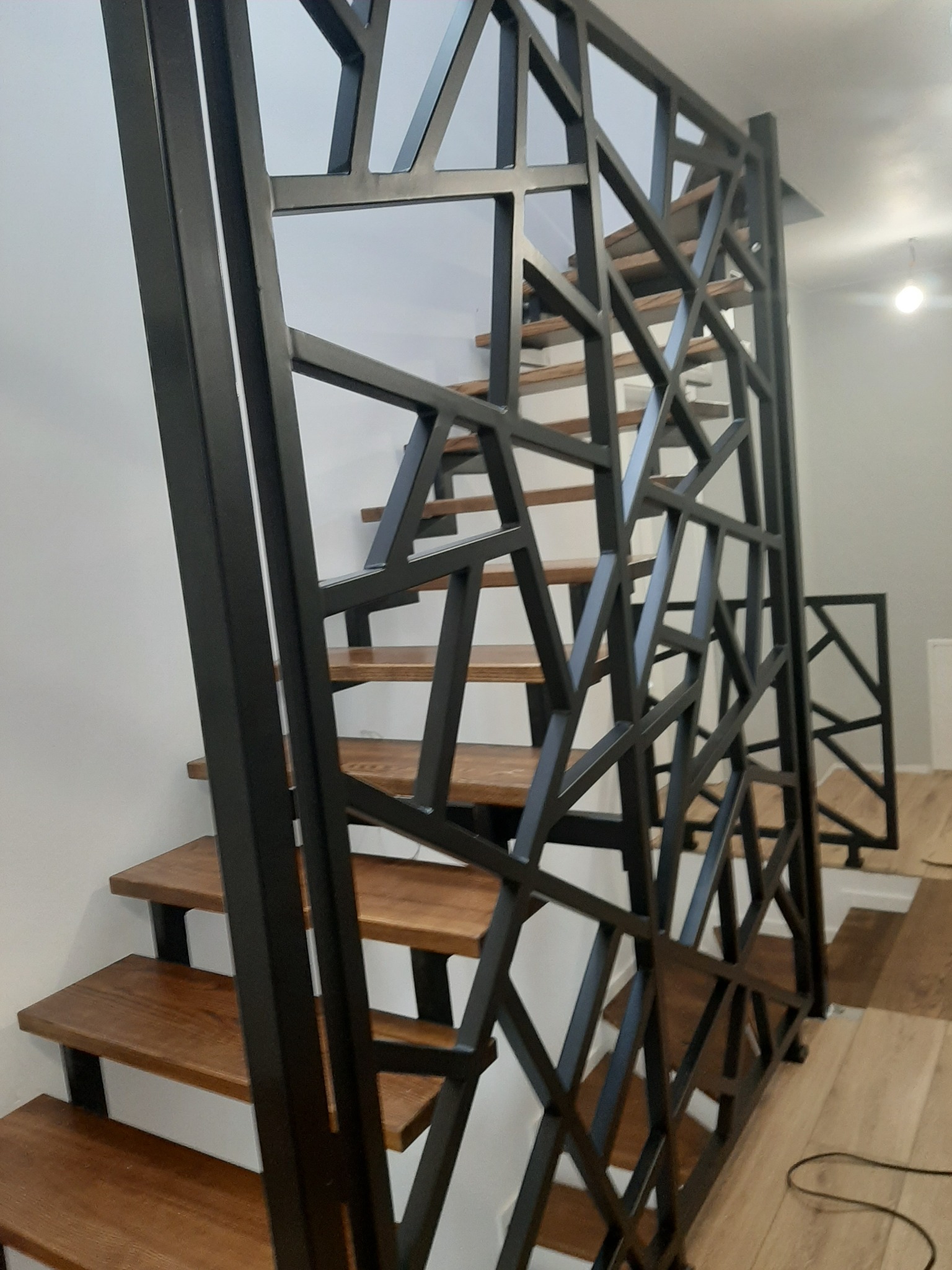 Nowoczesna klatka schodowa z drewnianymi stopniami i ażurową, geometryczną balustradą z czarnego metalu.