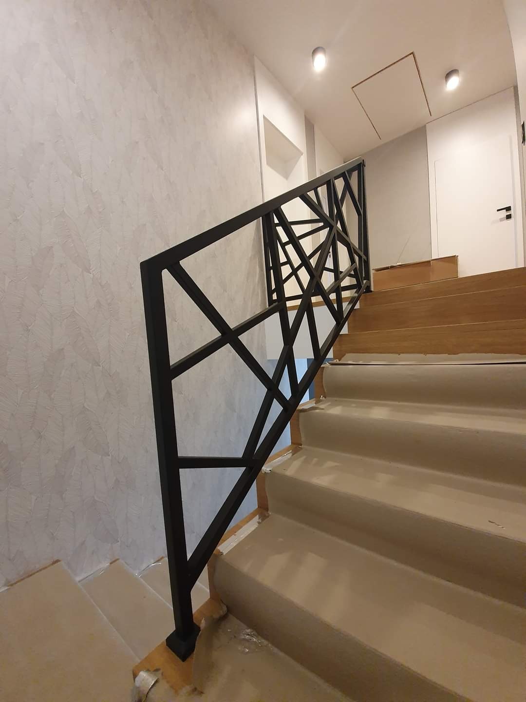 Nowoczesna balustrada metalowa o geometrycznym wzorze, czarna, zamontowana na schodach z drewnianymi stopniami i jasnymi podstopnicami, ściana z dekoracyjną fakturą, widoczne zabezpieczenia...