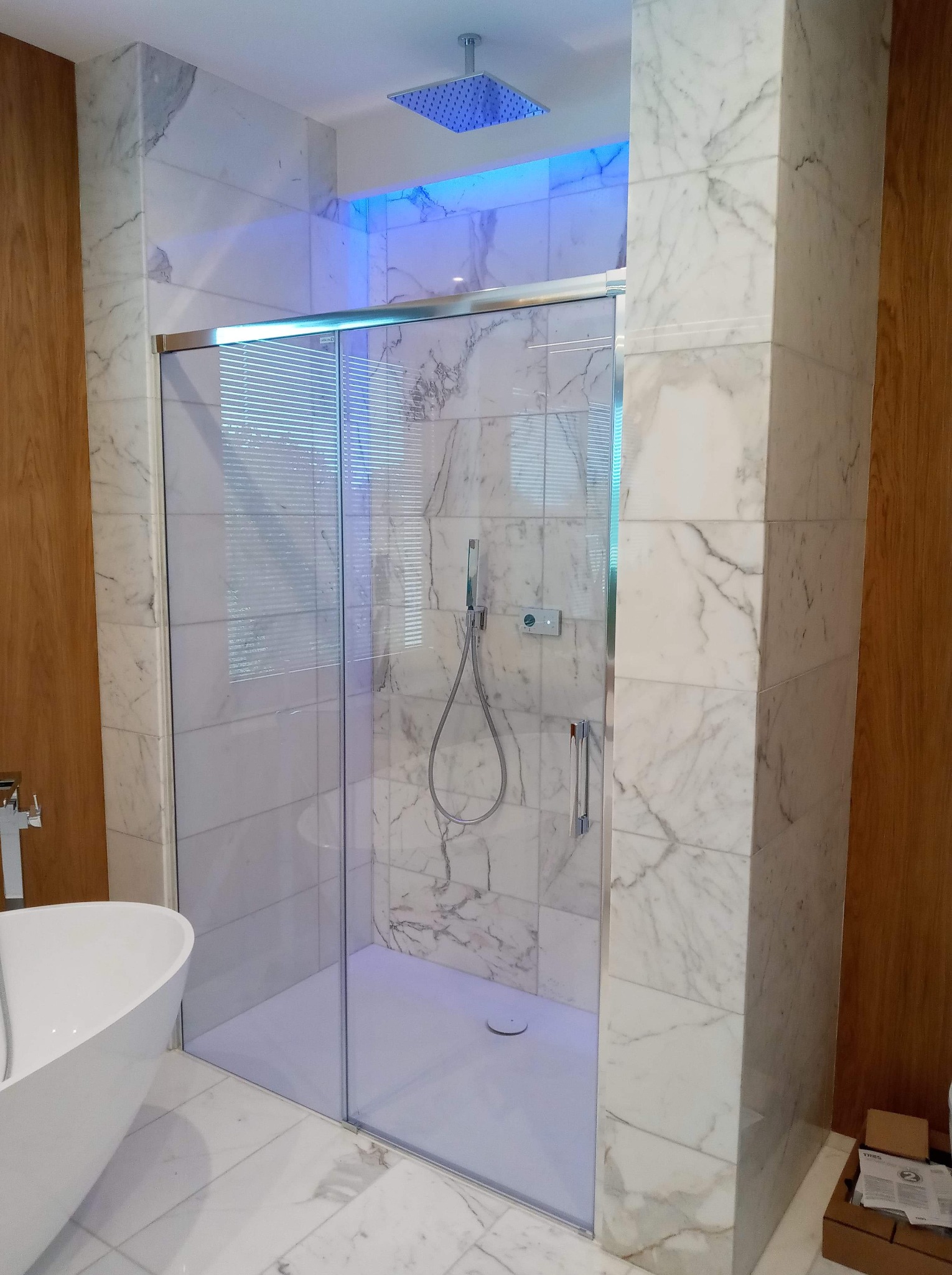 Nowoczesna łazienka z kabiną prysznicową typu walk-in, wykończona marmurem, z podświetlanym deszczownicą i białą, wolnostojącą wanną.