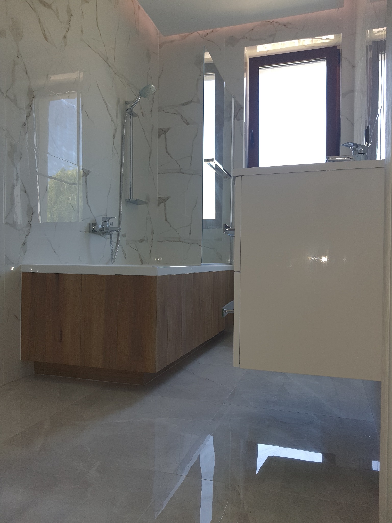 Nowoczesna łazienka z marmurowymi płytkami, drewnianą obudową wanny i białymi szafkami, okno z ciemną ramą w tle.