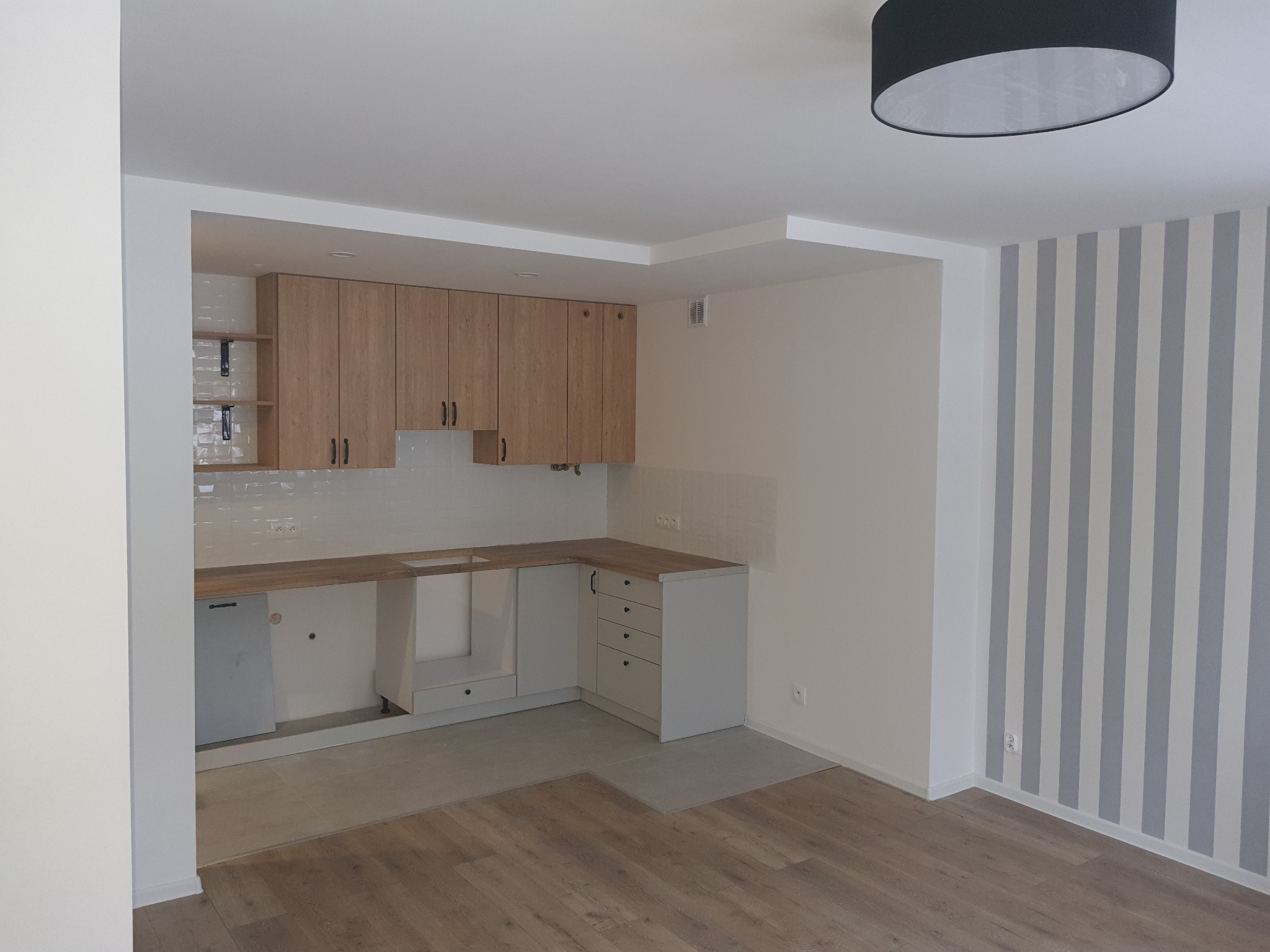 Wnętrze kuchni w trakcie remontu: widoczne niedokończone szafki kuchenne z drewnianymi frontami, białe płytki na ścianie, fragment podłogi z drewnianymi panelami oraz ściana w szaro-białe pasy.