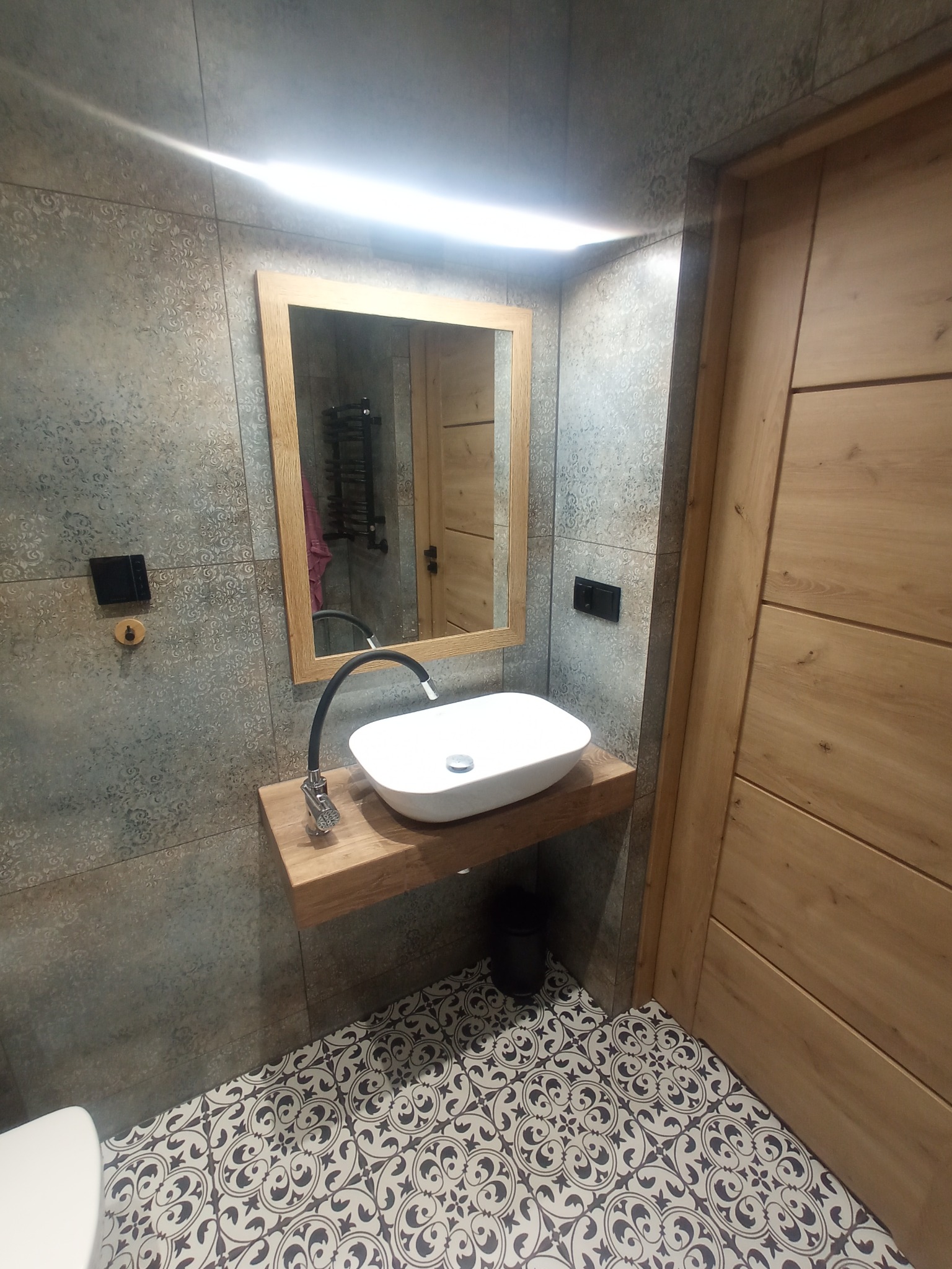 Umywalka nablatowa o owalnym kształcie z czarną, giętką baterią, zamontowana na drewnianym blacie w łazience wykończonej szarymi płytkami z delikatnym wzorem i podłogą w biało-czarne ornamenty.