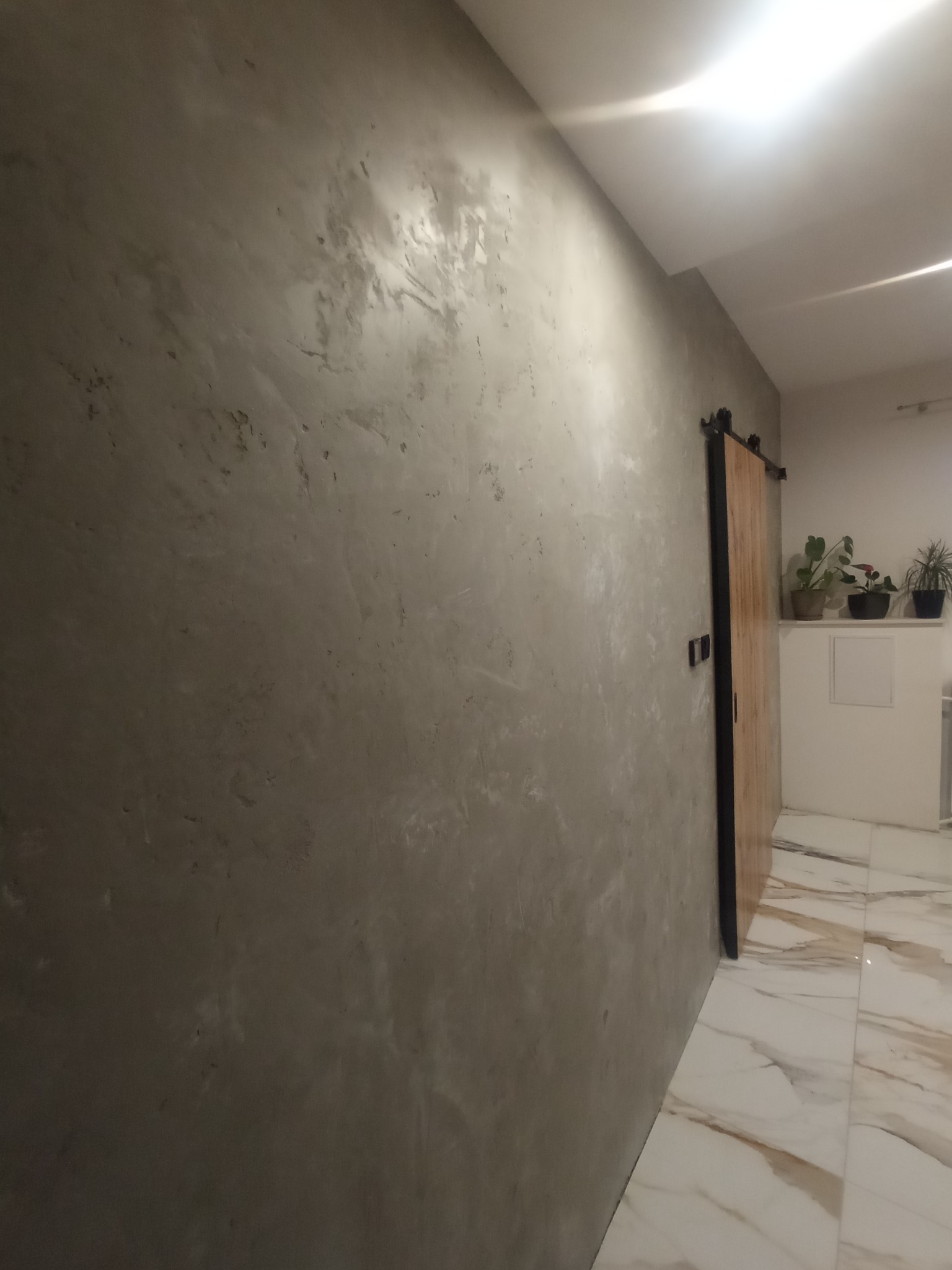 Ściana z szarą, dekoracyjną strukturą tynku w bliskim ujęciu, widoczna drewniana przesuwna brama z czarnym okuciem i fragment marmurowej podłogi.