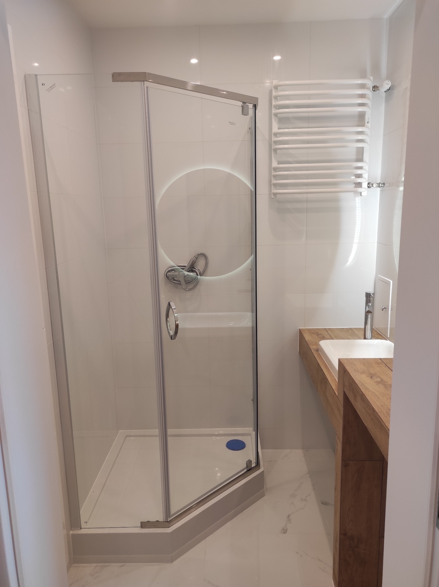 Nowoczesna łazienka z kabiną prysznicową z chromowanymi profilami, białymi płytkami, drewnianą umywalką nablatową i grzejnikiem drabinkowym.