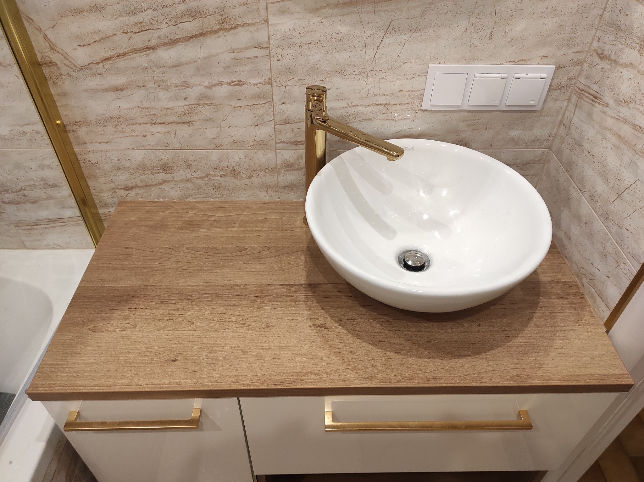 Biała, okrągła umywalka nablatowa z złotą baterią na szafce z blatem imitującym drewno, marmurowe płytki na ścianie i biały panel z włącznikami światła.