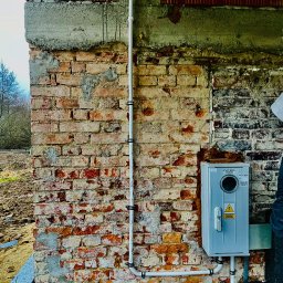 TendOO Electrics Filip Brzuchalski - Szara skrzynka elektryczna Inco Bex zamontowana na starej, częściowo otynkowanej ścianie z czerwonej cegły z widocznymi przewodami elektrycznymi. Na górze ściany widoczny surowy beton i fragmenty...