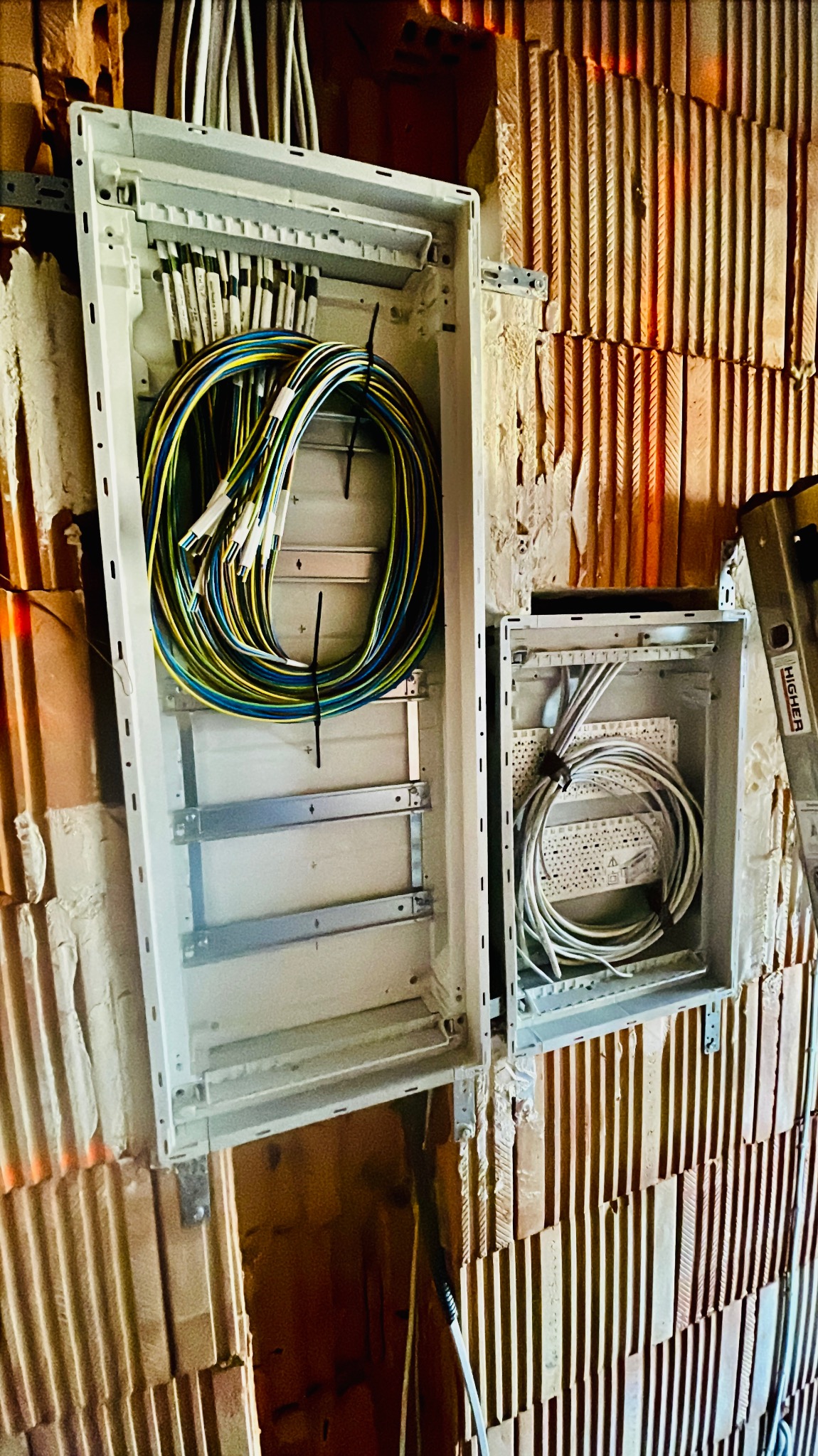 Dwie otwarte, białe skrzynki elektryczne zamontowane na ceglanej ścianie, z widocznymi kablami wewnątrz; większa skrzynka z kablami w kolorach niebieskim, żółtym i zielonym, mniejsza z białymi...