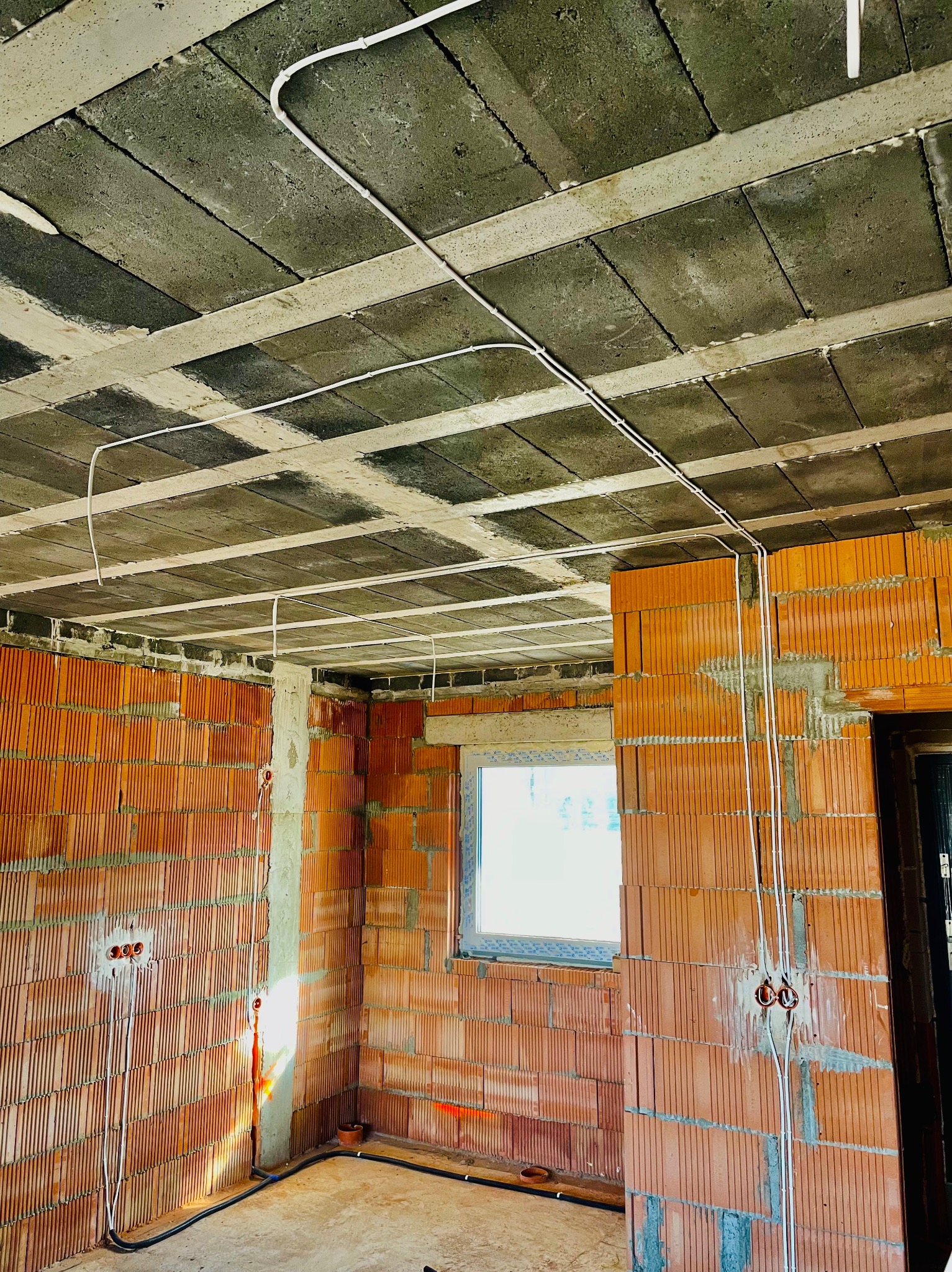 Wewnętrzne ściany z czerwonej cegły z widocznymi przewodami elektrycznymi, szary betonowy sufit z widocznymi spoinami, okno w tle.