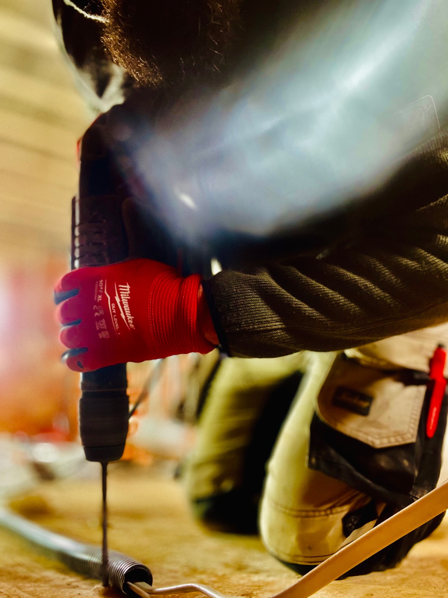 Pracownik w czerwonych rękawicach Milwaukee wierci otwór w metalowej sprężynie na drewnianej powierzchni, widoczny fragment spodni roboczych i ołówek stolarski.