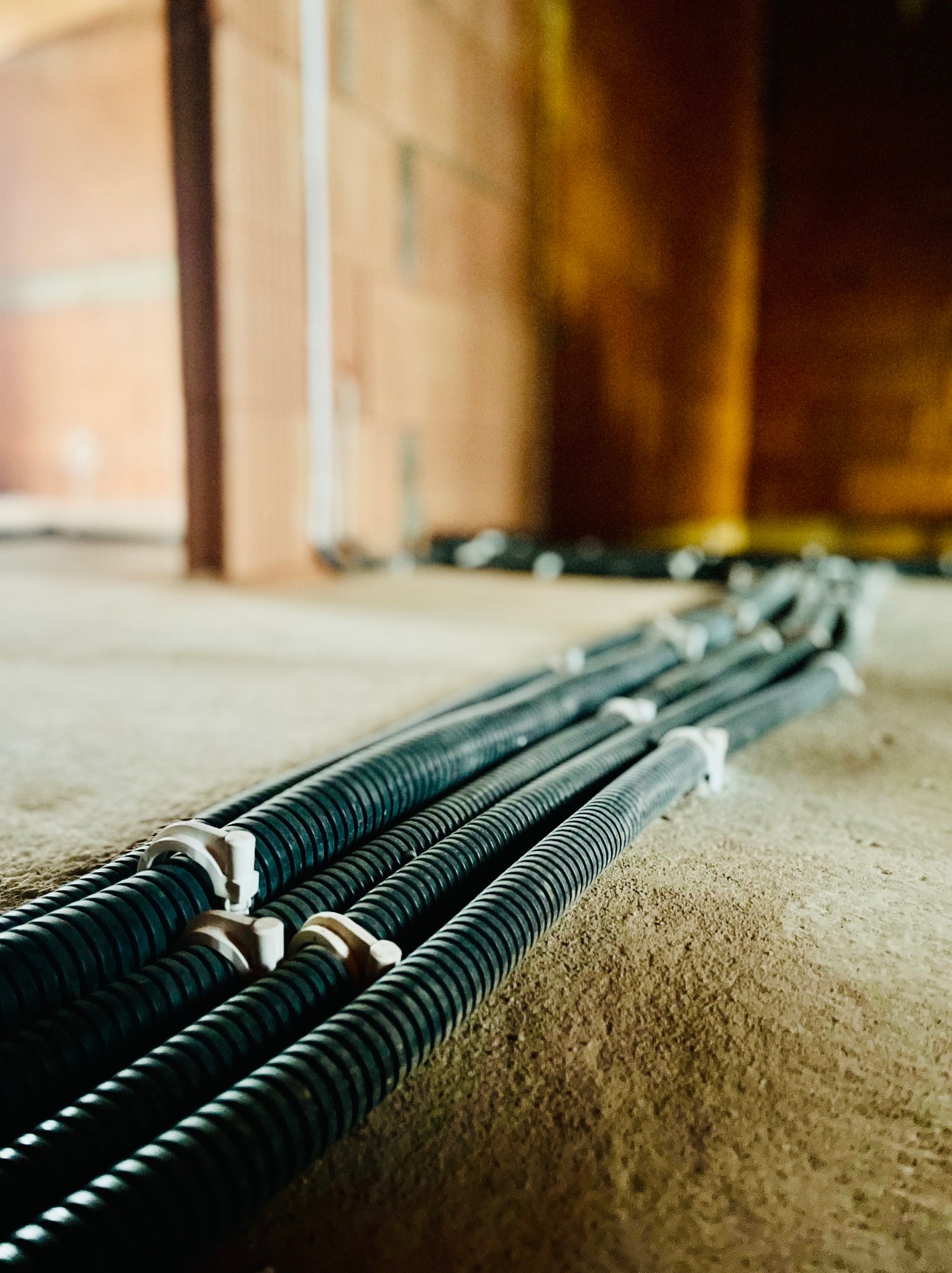 Rząd czarnych, karbowanych rur osłonowych na podłodze w trakcie budowy, zabezpieczonych białymi uchwytami, z widoczną ceglaną ścianą w tle.