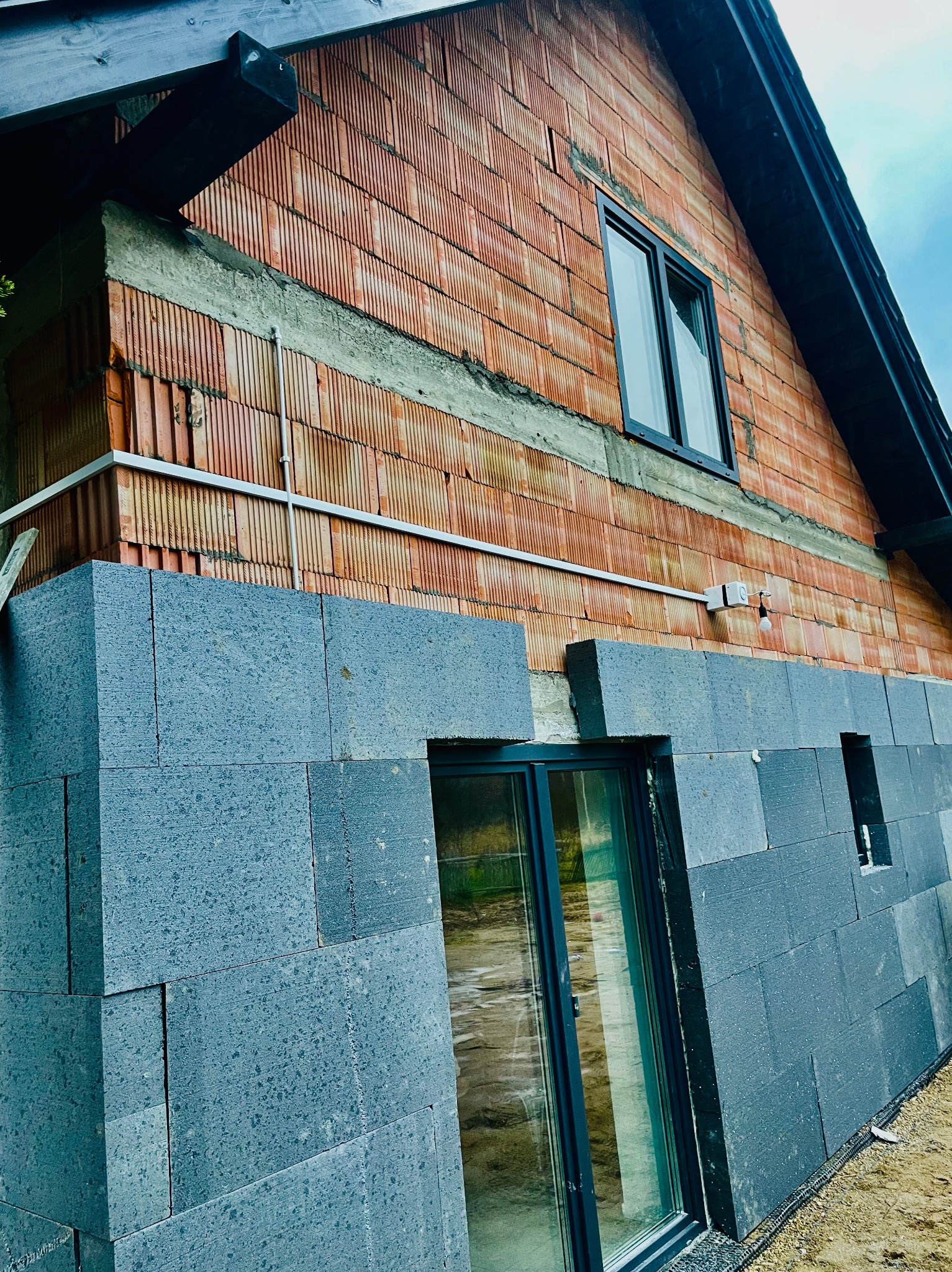 Surowy stan budynku z cegły ceramicznej i szarego betonu komórkowego z oknami w czarnych ramach, widoczne przewody elektryczne na elewacji.