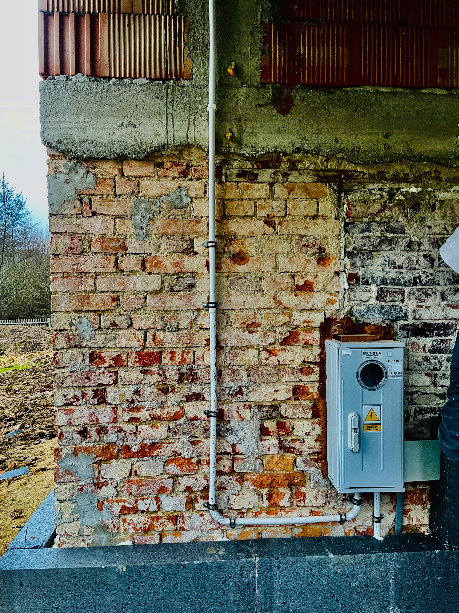 Szara skrzynka elektryczna Inco Bex zamontowana na starej, częściowo otynkowanej ścianie z czerwonej cegły z widocznymi przewodami elektrycznymi. Na górze ściany widoczny surowy beton i fragmenty...