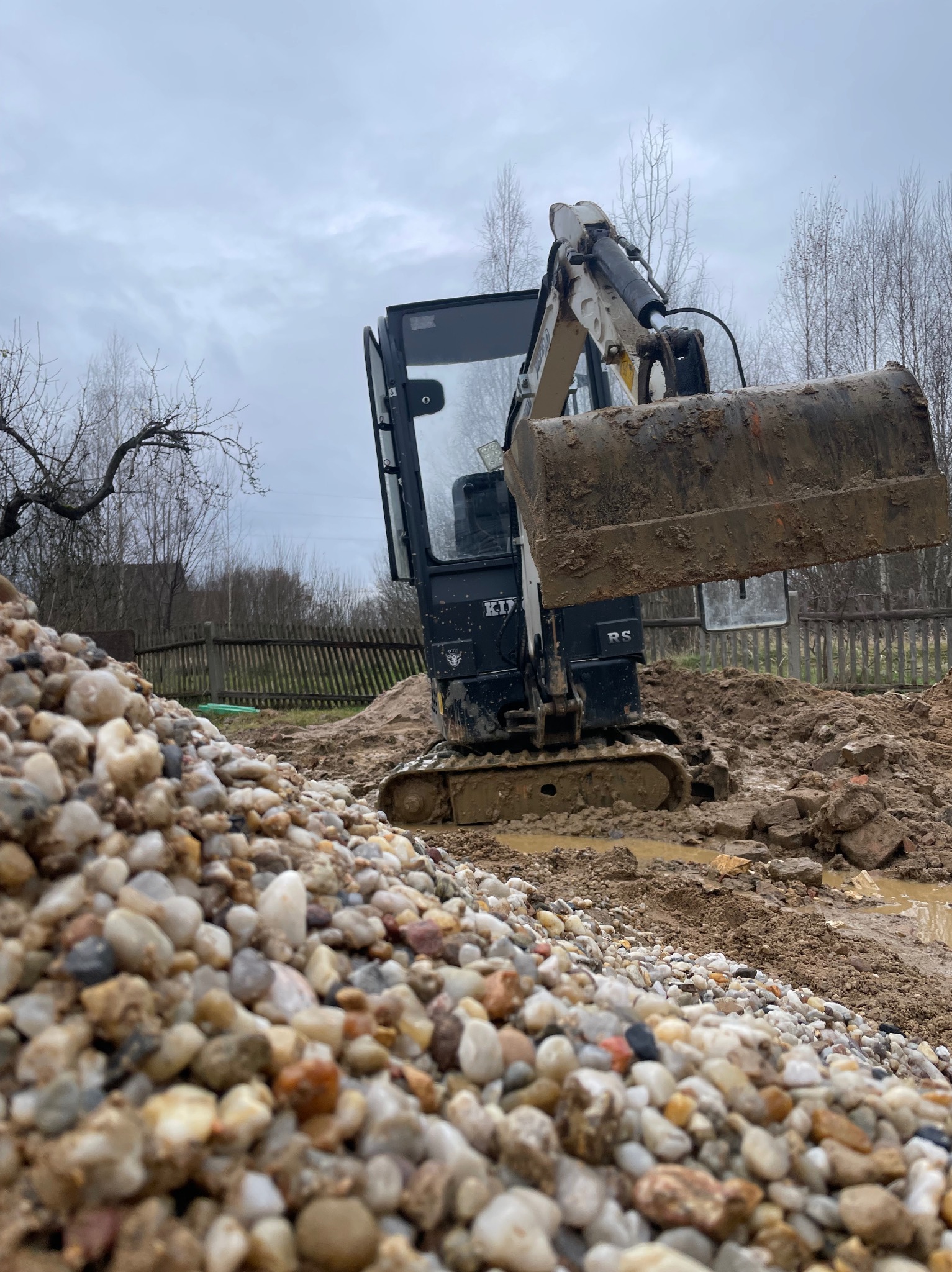 Mała koparka z łyżką pełną ziemi na błotnistym placu budowy, w tle sterta kamieni i drewniany płot.