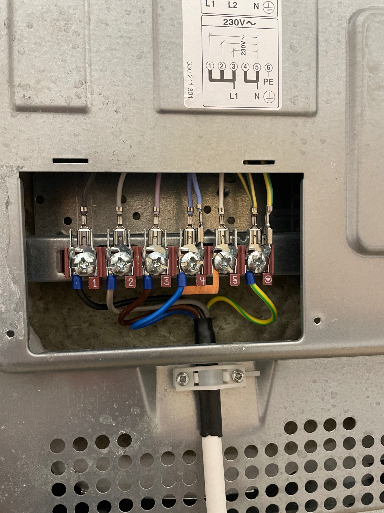 Szczegółowe ujęcie tylnej części płyty indukcyjnej z widocznymi podłączeniami elektrycznymi: przewody w kolorach białym, brązowym, niebieskim, fioletowym i żółto-zielonym podłączone do listwy...