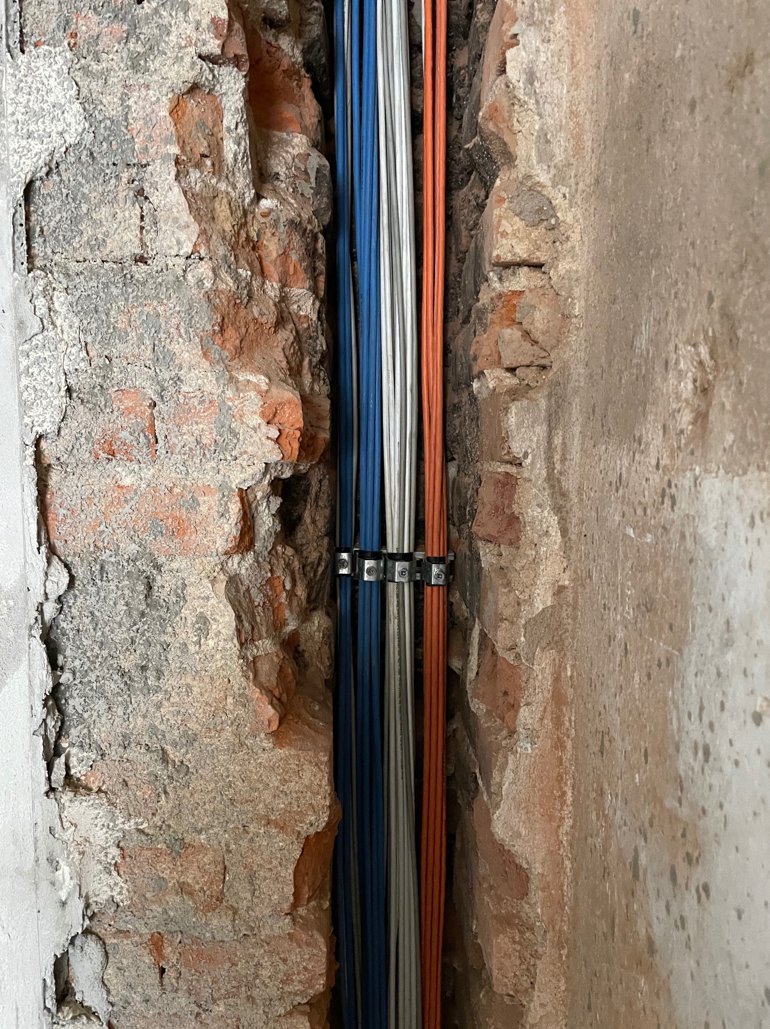 Wiązka kabli elektrycznych w kolorach niebieskim i pomarańczowym, przymocowana metalowymi uchwytami do bruzdy w ścianie z cegły i betonu.