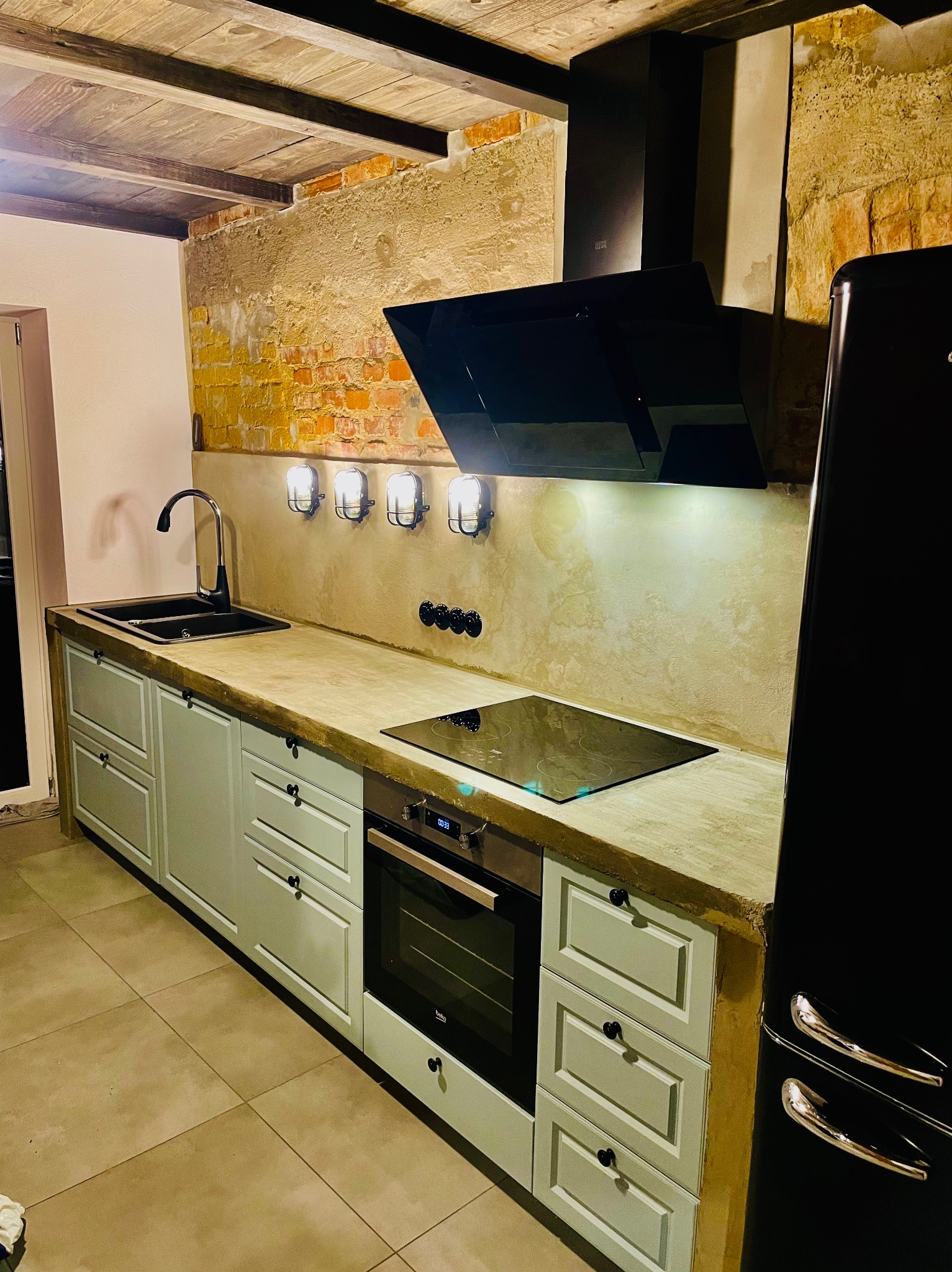 Kuchnia w stylu rustykalnym z szarymi szafkami, betonowym blatem, czarnym zlewem i baterią, płytą indukcyjną, piekarnikiem oraz czarną lodówką retro. Na ścianie widoczna cegła i lampy w stylu...