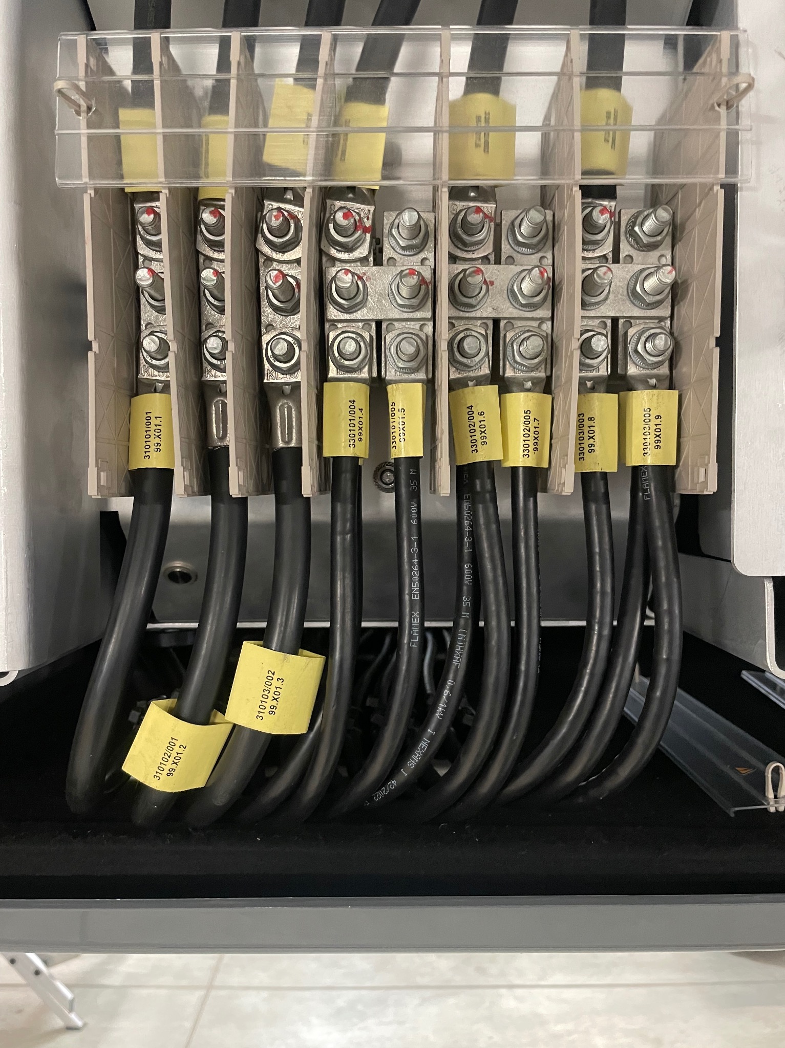 Szczegółowe ujęcie skrzynki elektrycznej z podłączonymi przewodami oznaczonymi żółtymi etykietami z numerami identyfikacyjnymi, widoczne śruby i elementy mocujące, całość za przezroczystą osłoną.